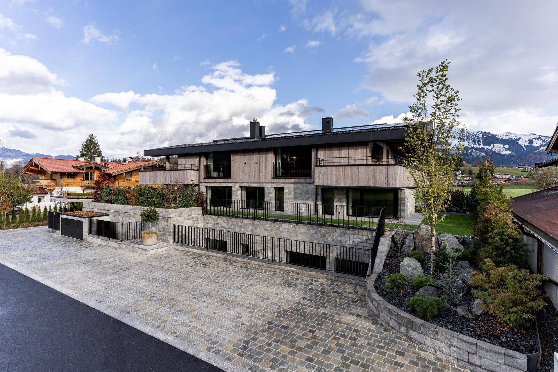 Moderne Architektur mit Holz- und Steinfassade, großzügiger gepflasterter Auffahrt und Bergblick.