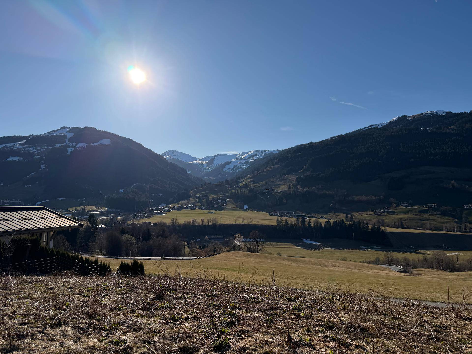 Sonnige Landschaft mit weiten Feldern, bewaldeten Hügeln und schneebedeckten Bergen im Hintergrund.