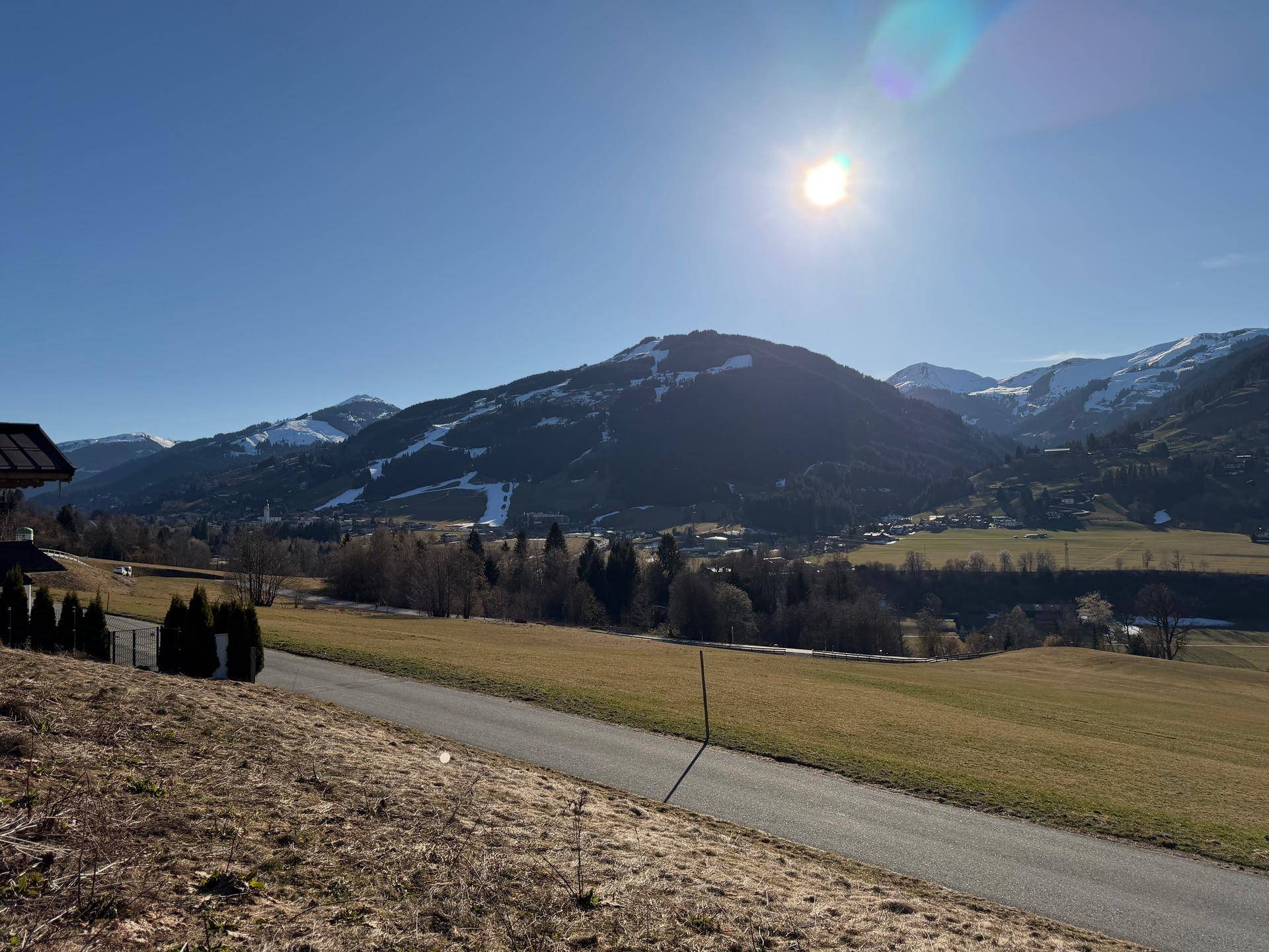 Ländliche Straße führt durch grüne Felder mit Blick auf bewaldete Hügel und schneebedeckte Berge.