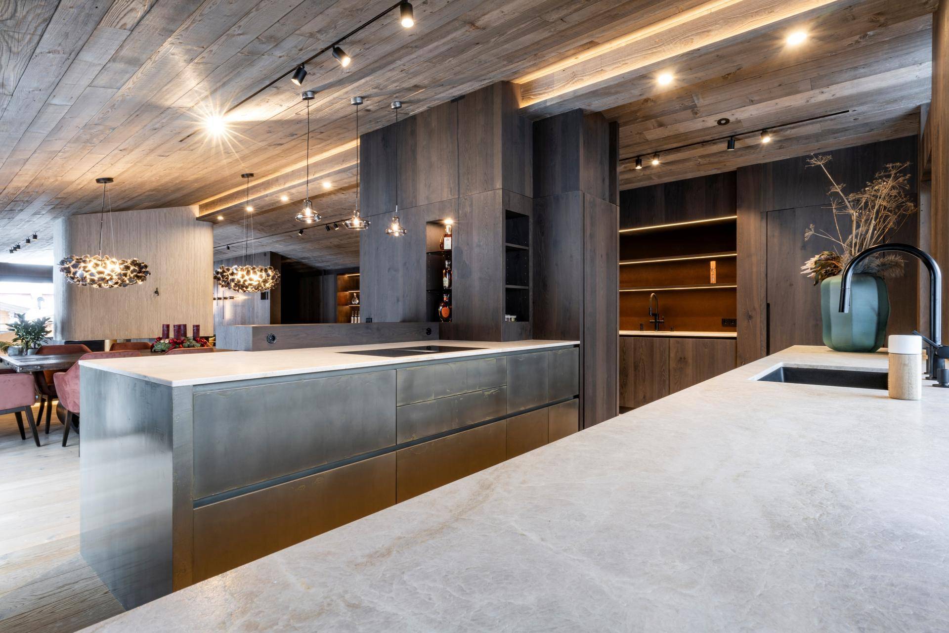 Offene, moderne Küche mit Holzdecken, Kücheninsel und hochwertigen Einbaugeräten in dunklem Holzdesign.