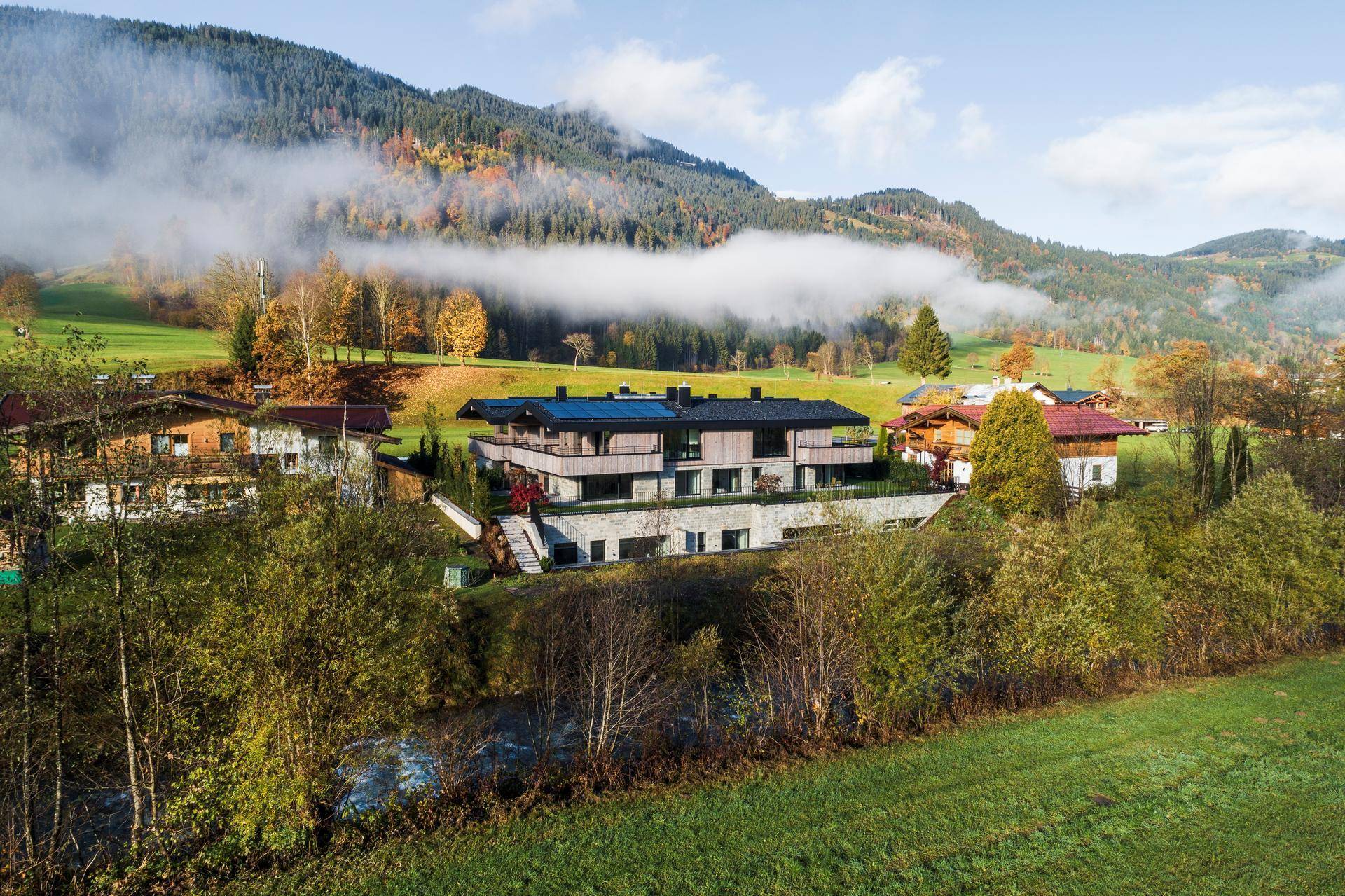 Modernes Gebäude mit Holzfassade und Photovoltaikmodulen, eingebettet in eine grüne Hügellandschaft mit Nebel.