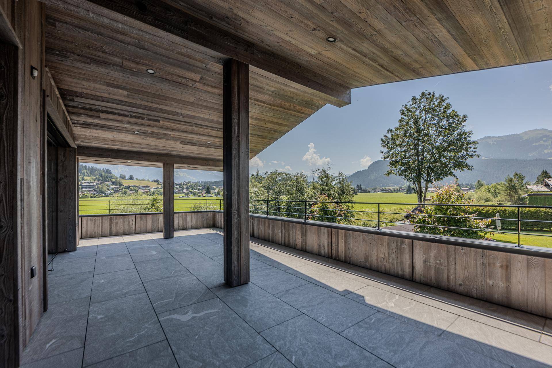 Große überdachte Terrasse mit Fliesenboden und weitem Blick auf die grüne Landschaft und Berge.