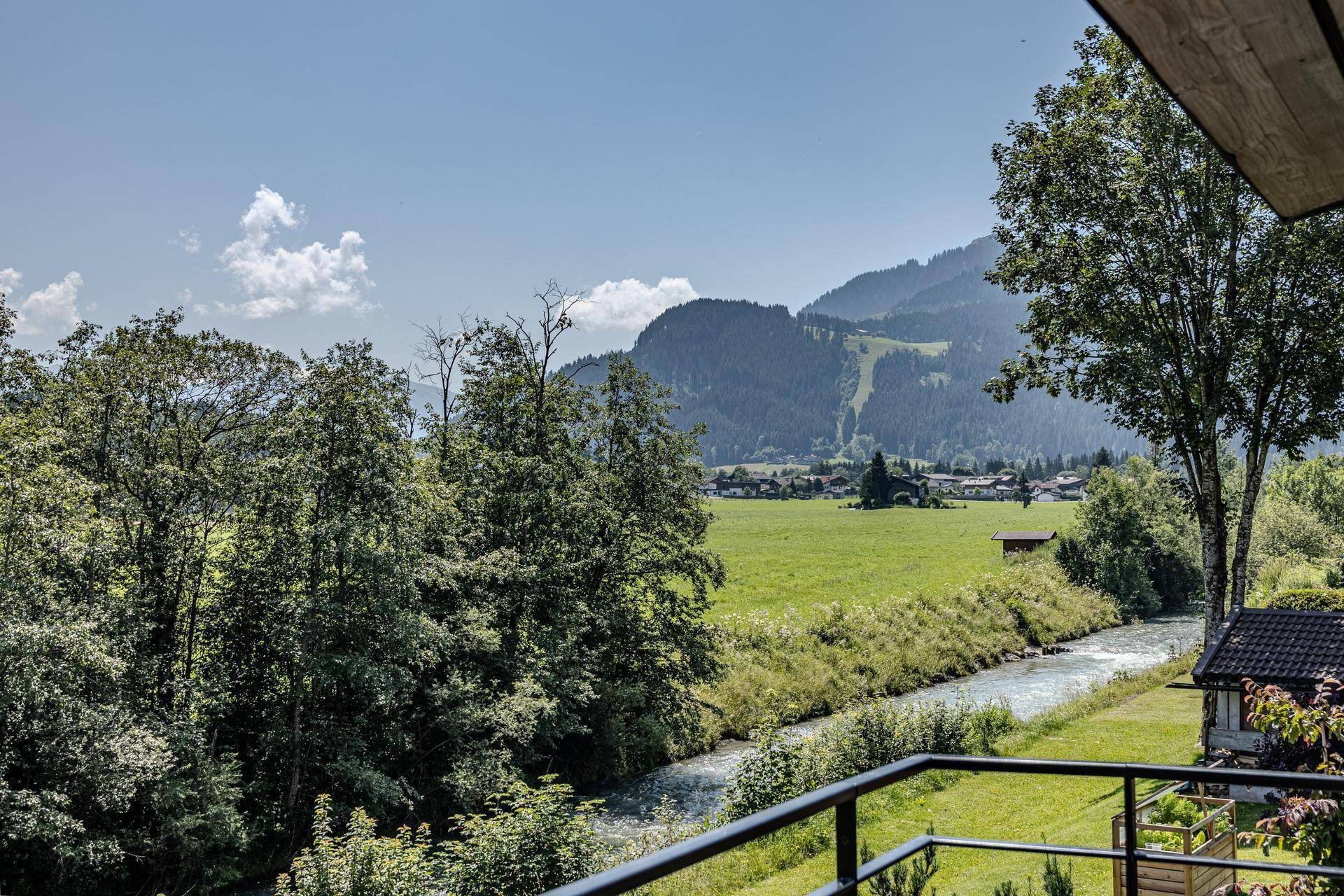 Panoramablick von einem Balkon auf eine grüne Wiese, einen Fluss und bewaldete Berge unter blauem Himmel.