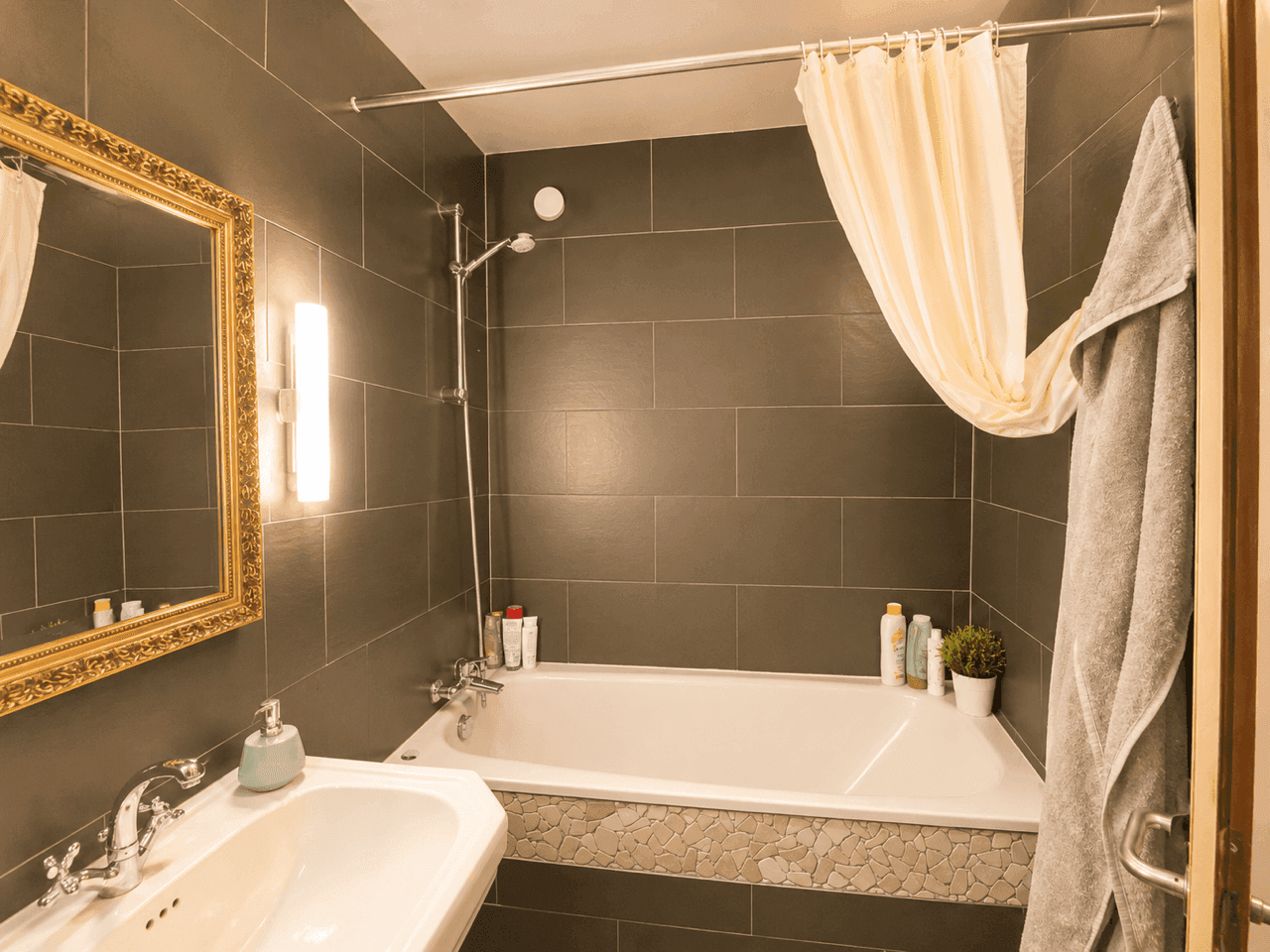 Badezimmer mit dunklen Fliesen, Badewanne mit Duschvorhang und Waschtisch.