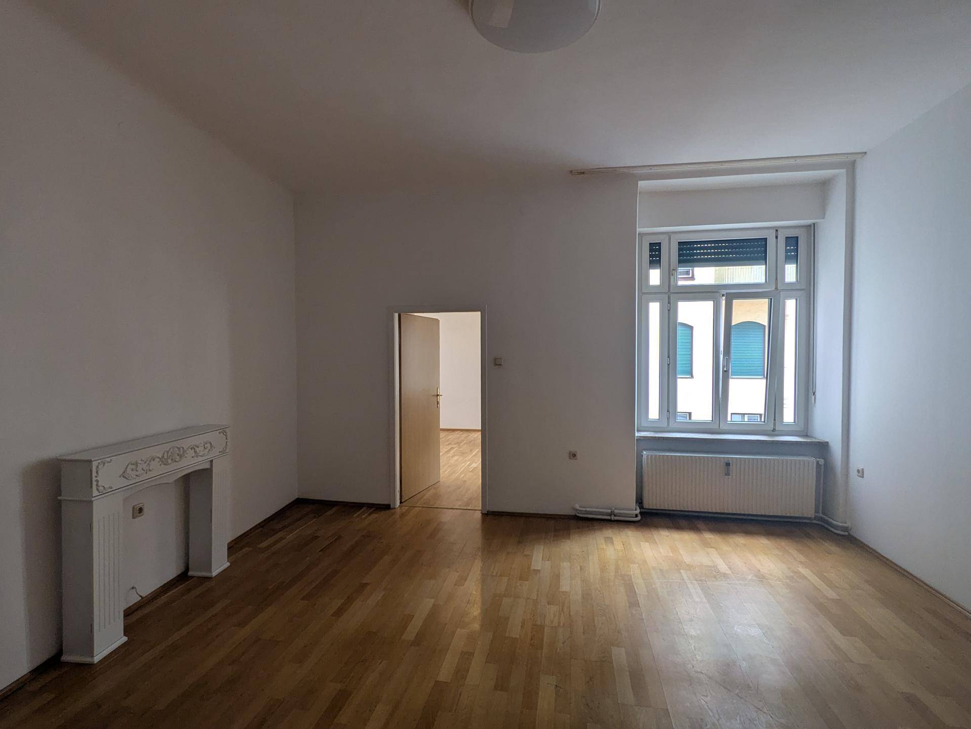 Geräumiges Wohnzimmer mit hellem Holzboden, dekorativem Kamin und großem Fenster mit Rollladen.