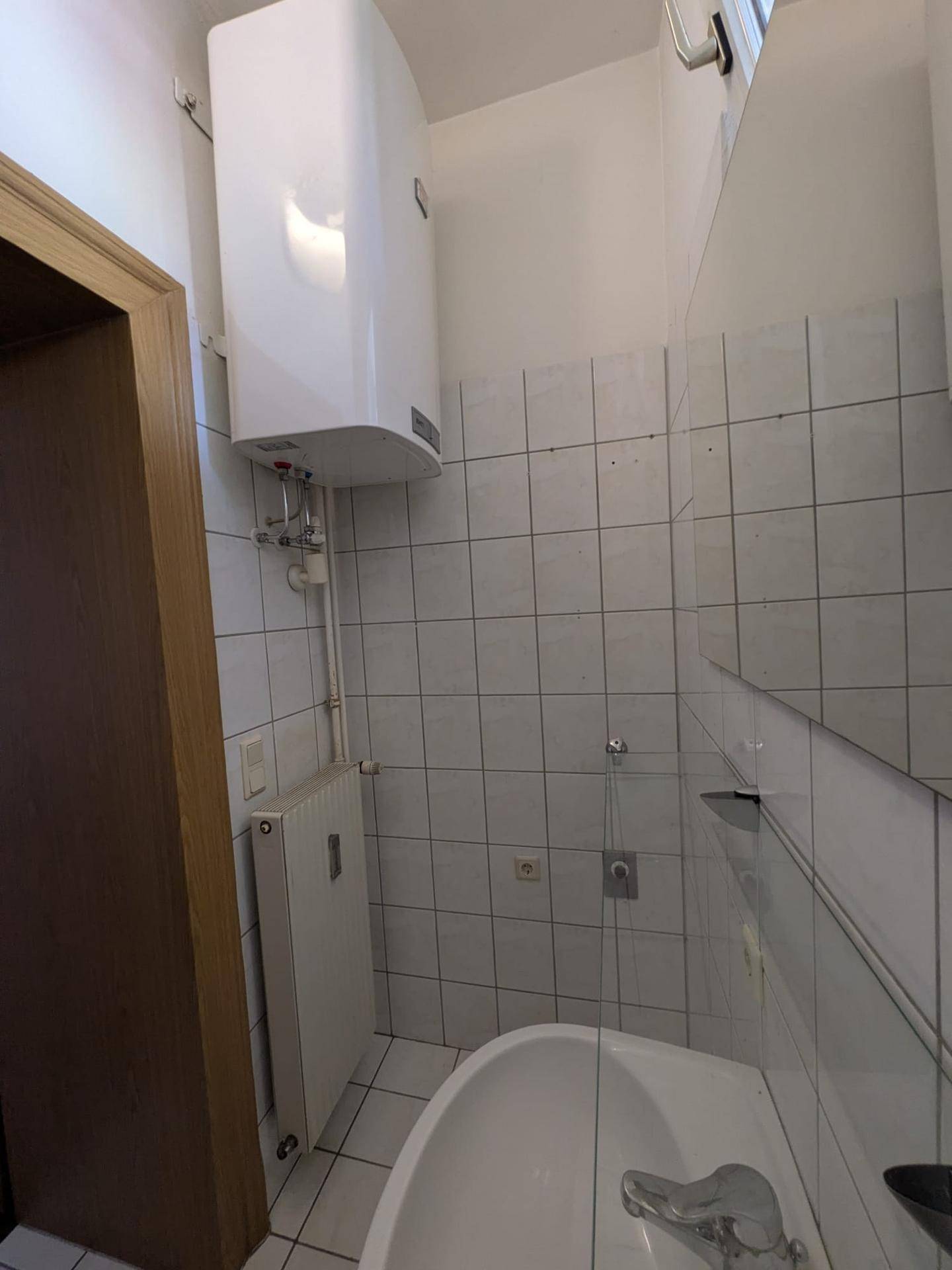 Badezimmer mit weißen Fliesen, Warmwasserboiler und Heizkörper, das eine grundlegende Ausstattung bietet.