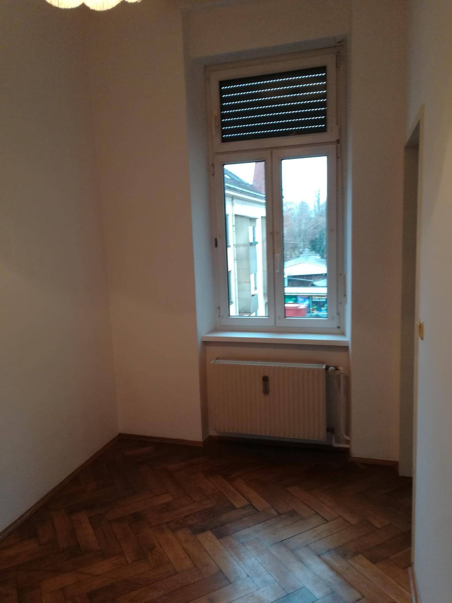 Helles Schlafzimmer mit Fischgrätparkett, Fenster mit Rollladen und Heizkörper, ideal für Ruhe und Entspannung.