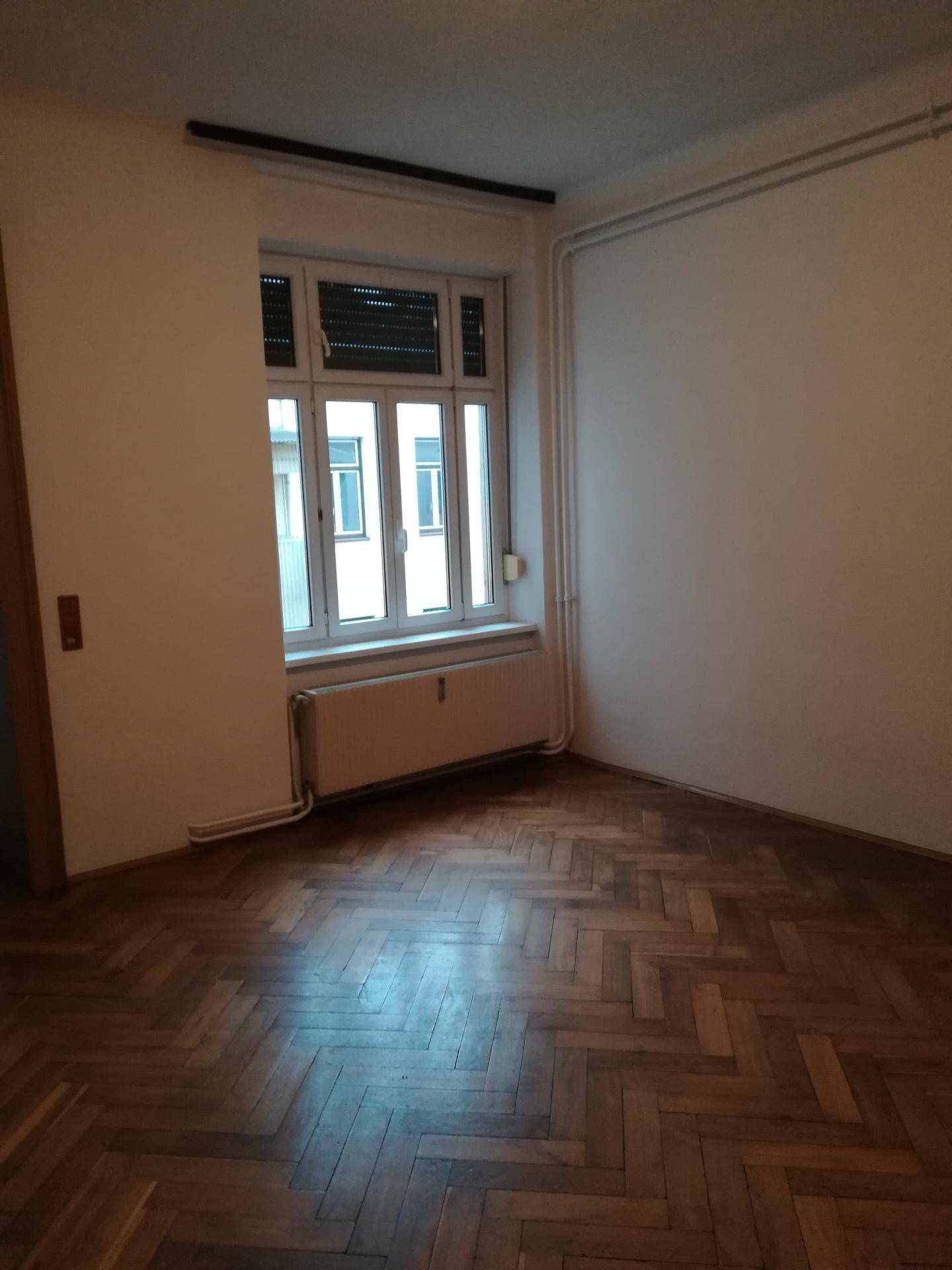 Gemütliches Zimmer mit klassischem Fischgrätparkett, großem Fenster und Heizkörper, das viel Charme bietet.