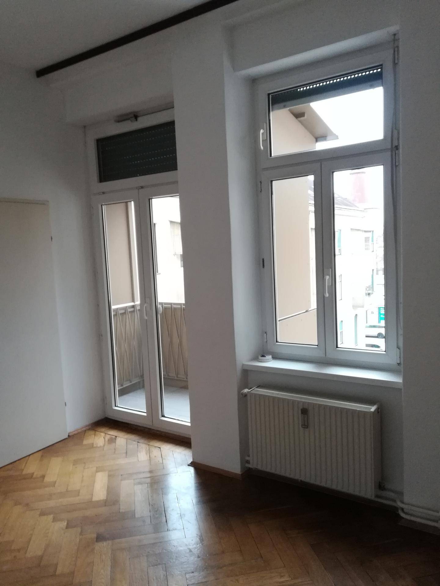 Heller Raum mit Fischgrätparkett und Zugang zu einem kleinen Balkon, der zusätzlichen Wohnraum im Freien bietet.