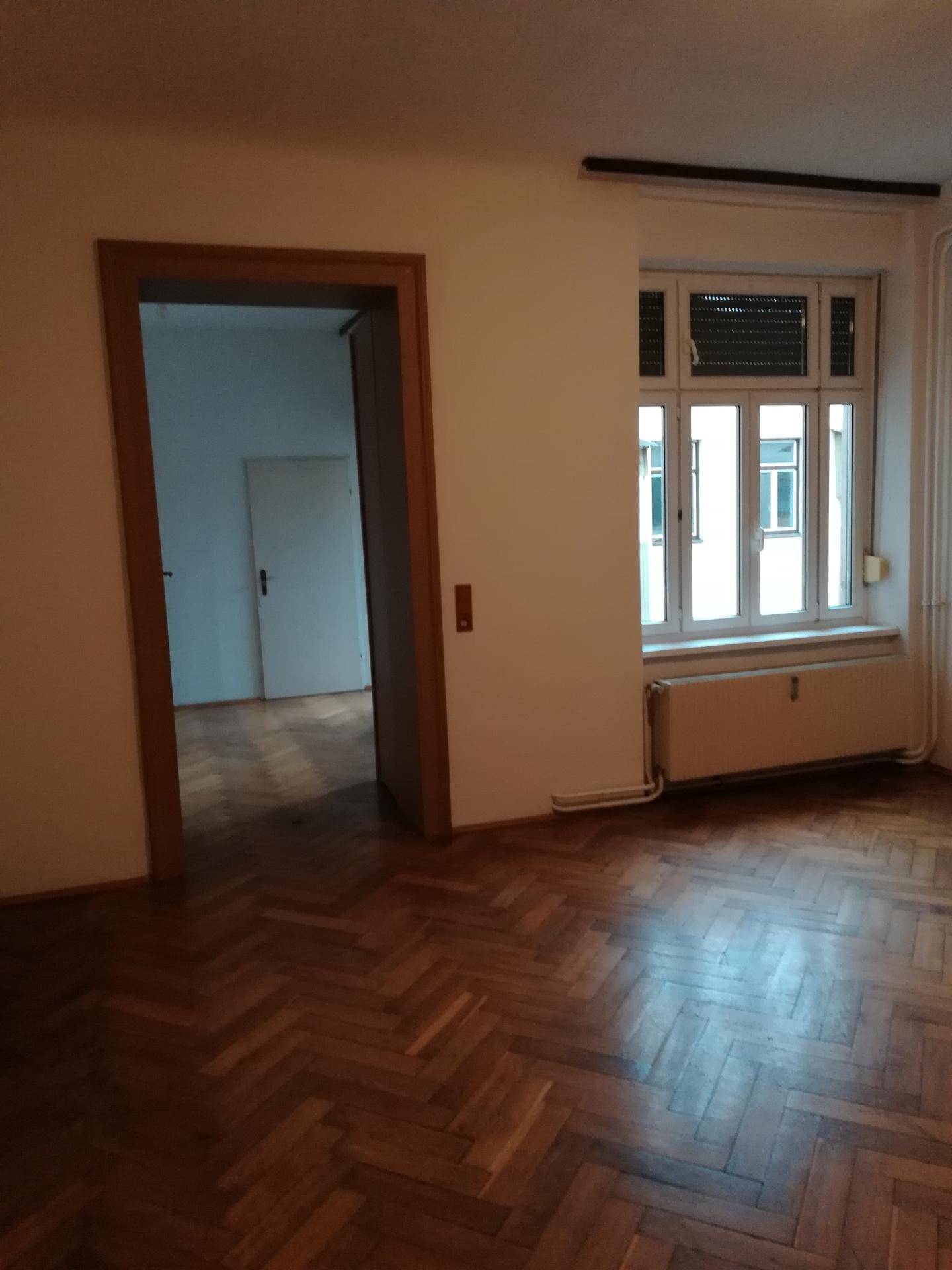 Wohnraum mit Fischgrätparkett und Fenster, der offen zu einem weiteren Zimmer führt und vielseitig nutzbar ist.