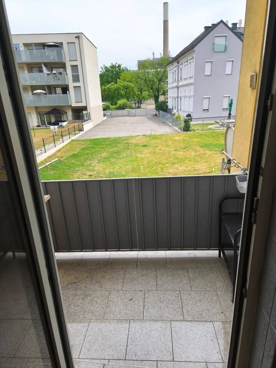 Blick vom Balkon auf eine Grünfläche und umliegende Gebäude mit Schornstein im Hintergrund.
