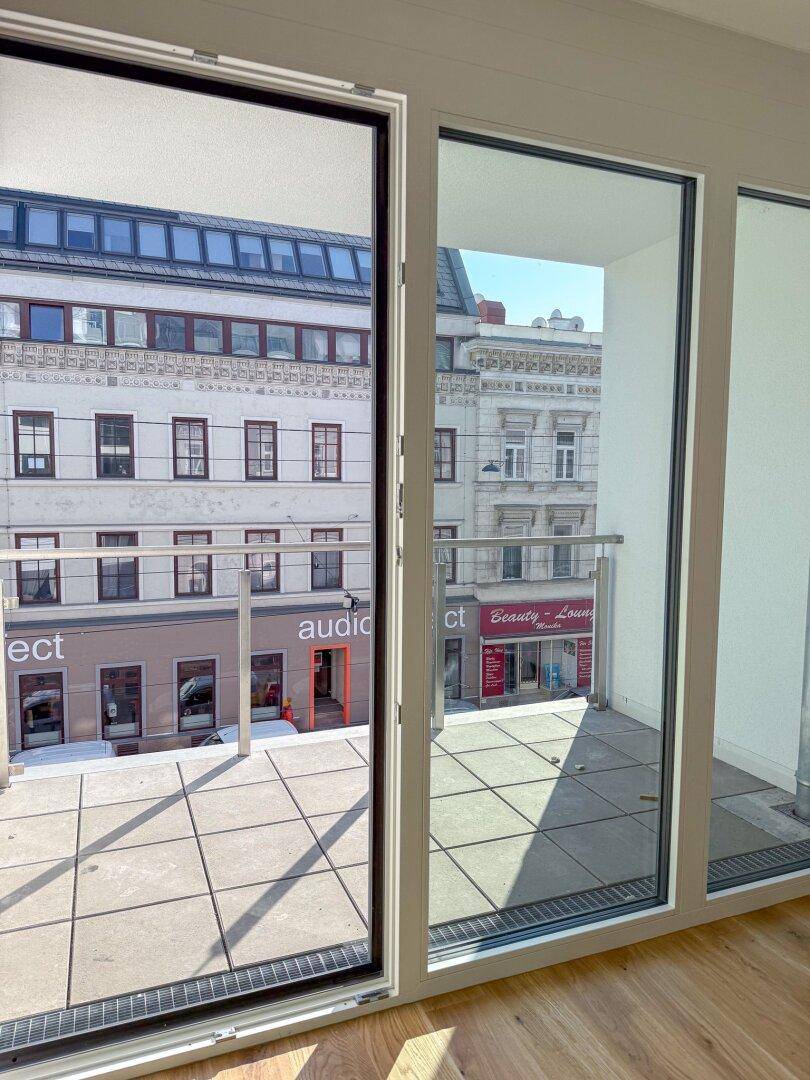 Blick von einem Innenraum mit Holzfußboden auf einen gefliesten Balkon und die belebte Stadtstraße.