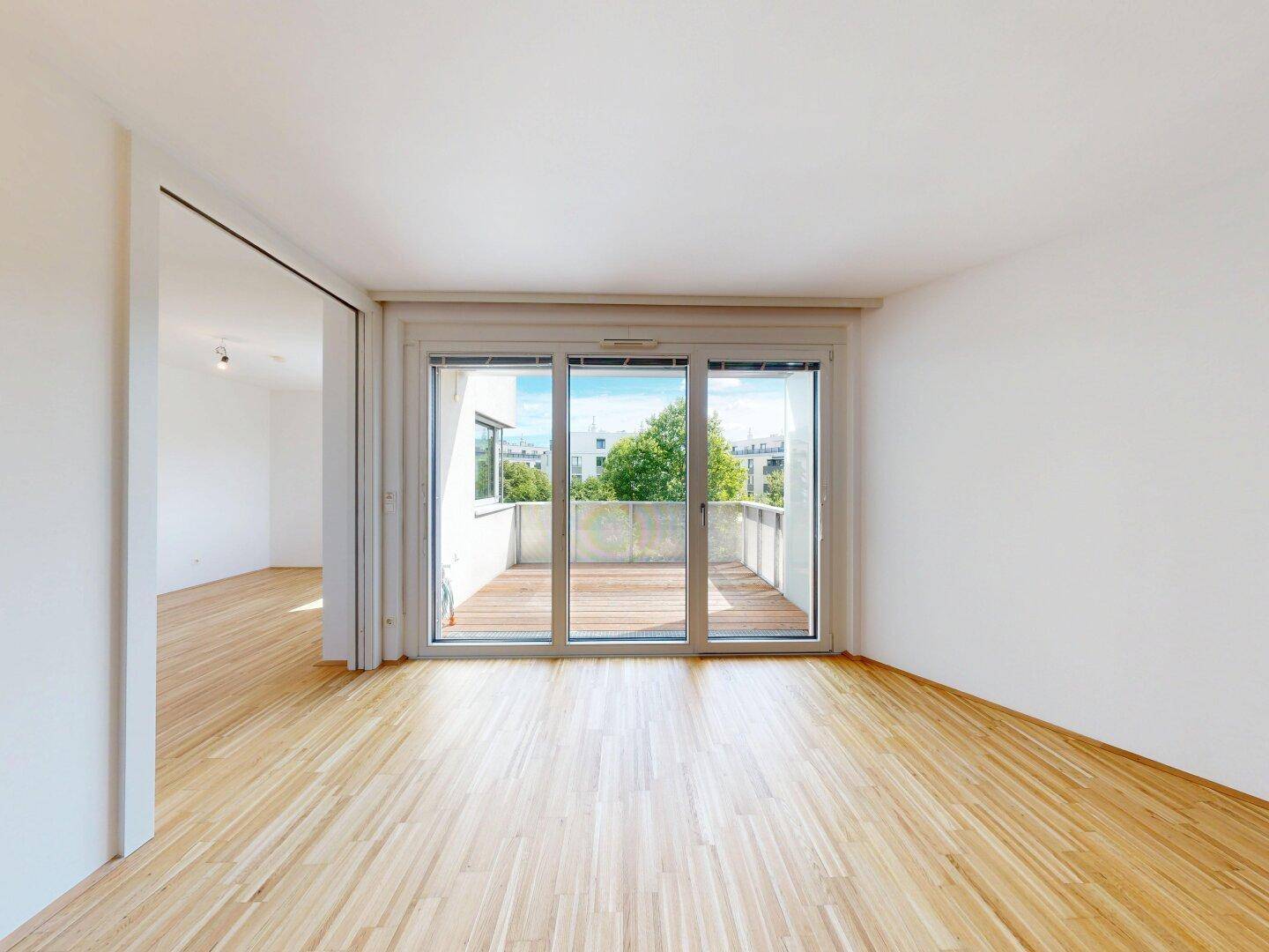 Geräumiger, heller Raum mit Holzboden und großen Schiebetüren, die zum Balkon führen.