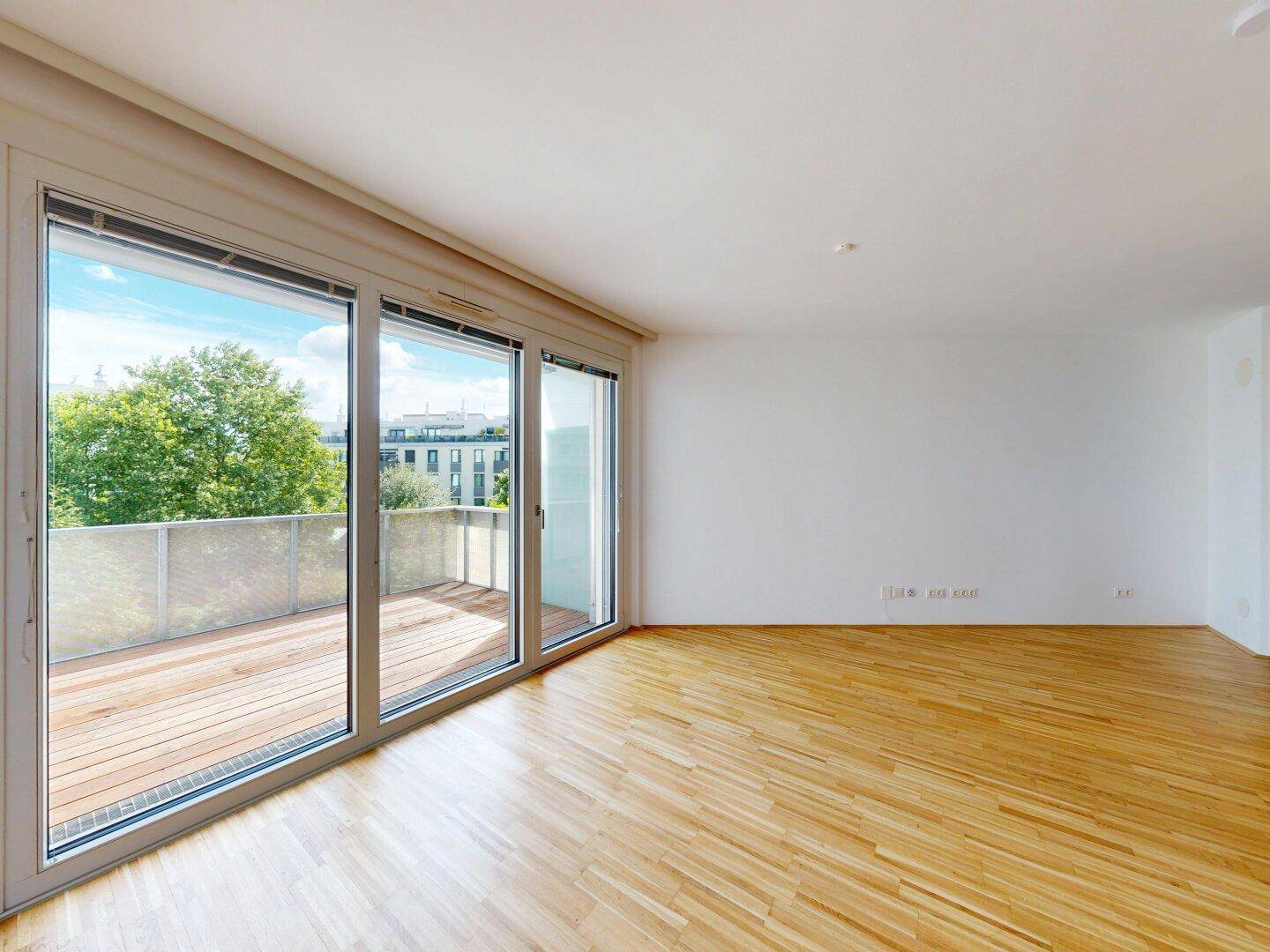 Heller Wohnbereich mit Holzboden und breiter Fensterfront, die viel Tageslicht hereinlässt.