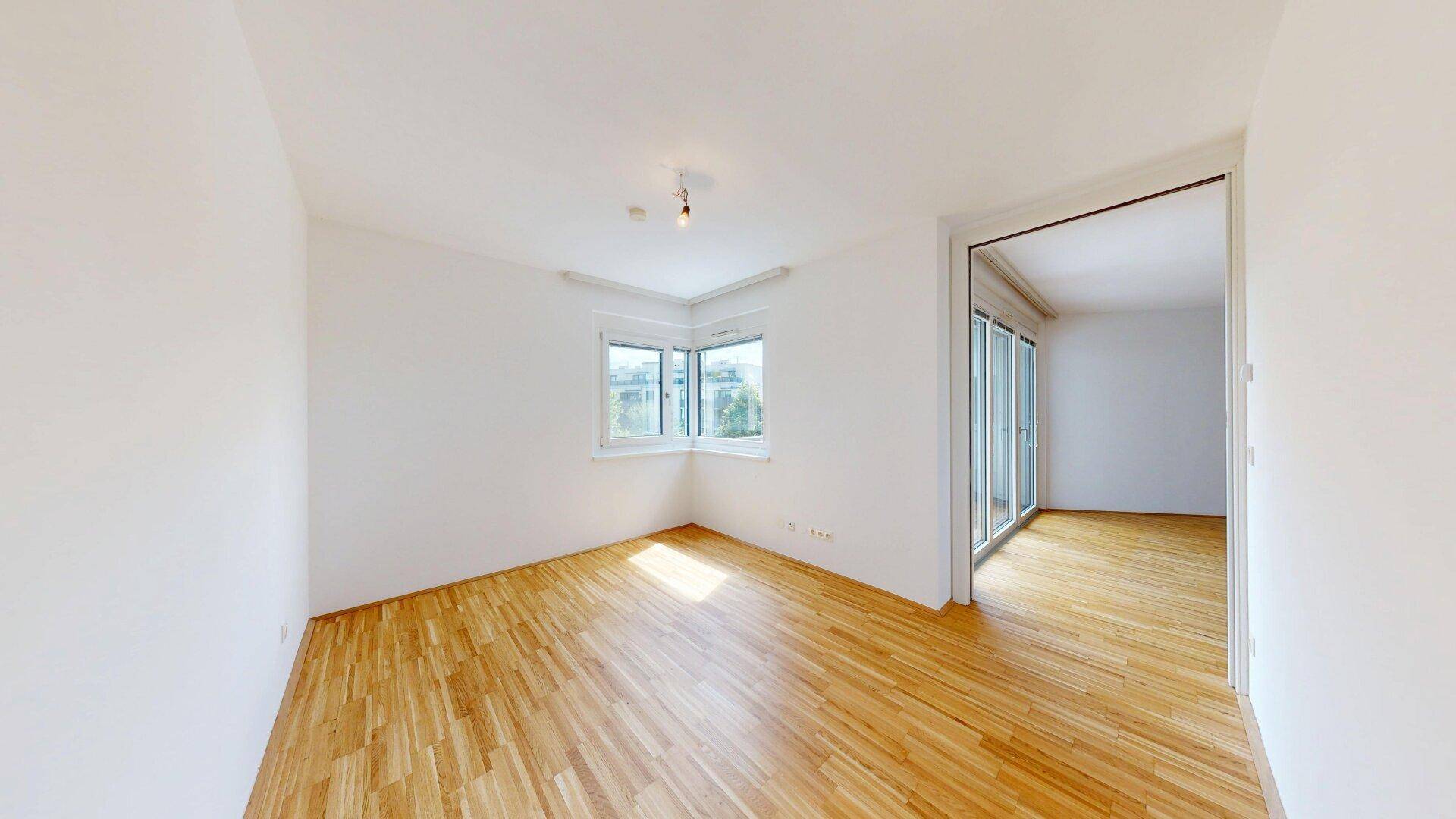 Heller, leerer Raum mit Holzboden und Eckfenster, bietet viel Tageslicht und Ausblick.
