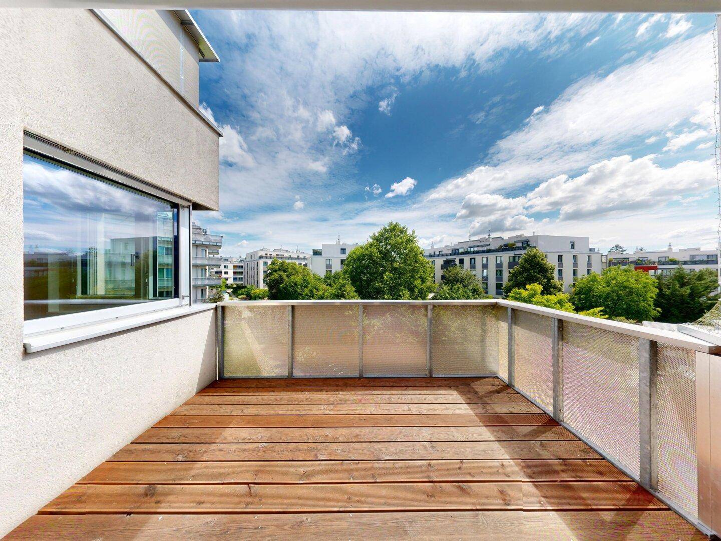 Heller Balkon mit Holzboden und Blick auf die umliegende grüne Umgebung und Gebäude.