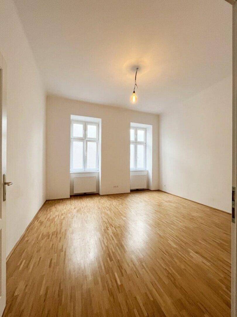 Leeres Zimmer mit hellem Parkettboden, weißen Wänden und zwei großen Fenstern.