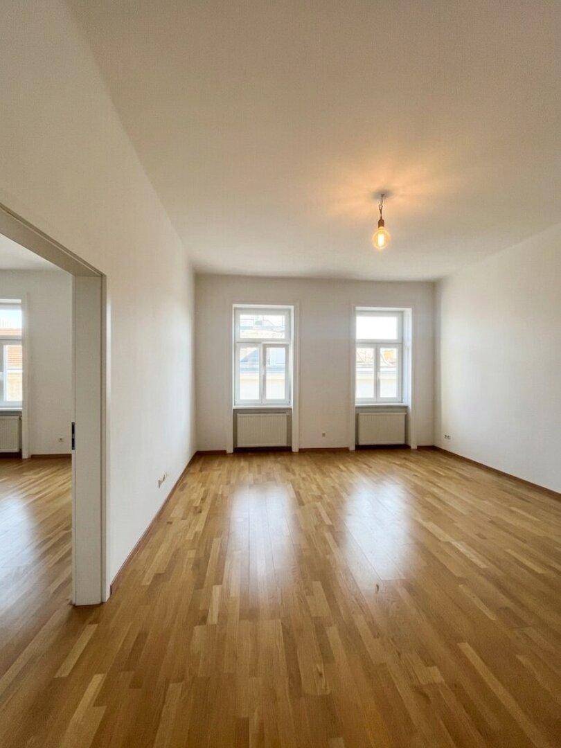 Großer, heller Raum mit Parkettboden und zwei Fenstern, ideal für Wohnbereich.