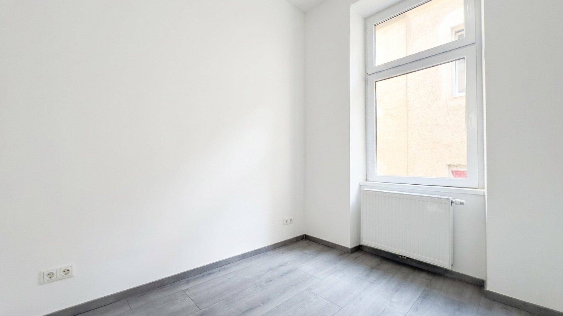 Kleiner, heller Raum mit einem Fenster und grauem Laminatboden.