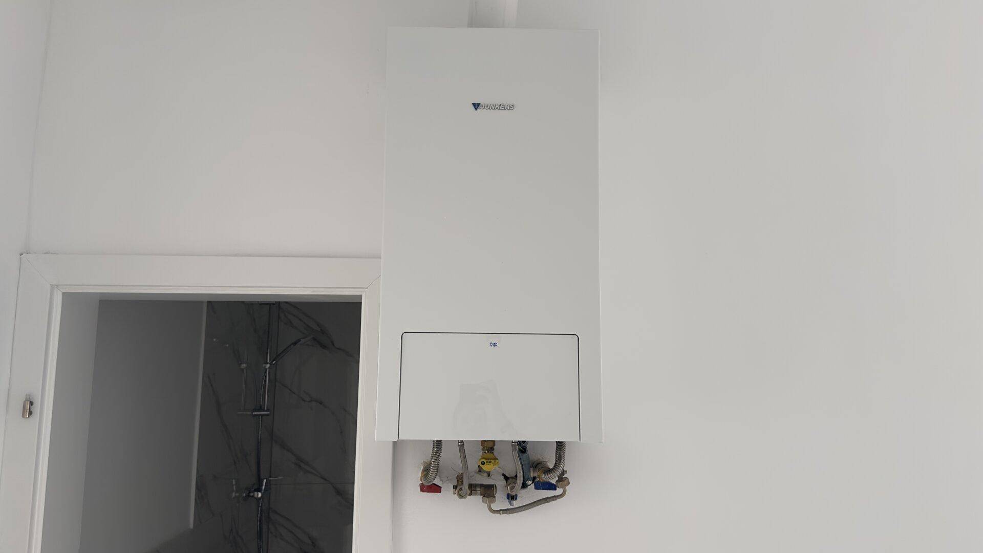 Weißer Heizkessel an einer schlichten weißen Wand, mit sichtbaren Anschlüssen und offener Tür.