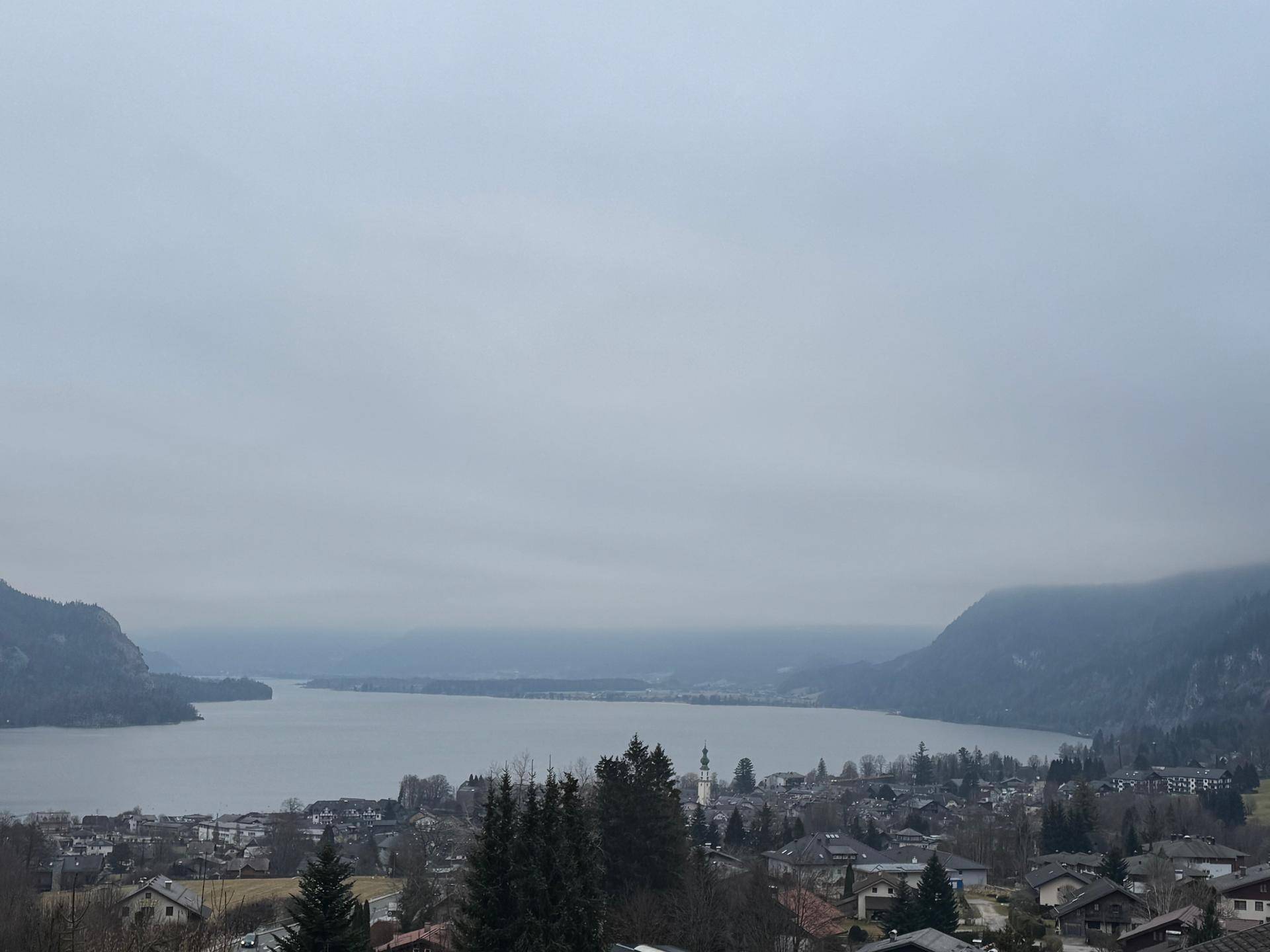 Weitläufiger Blick über einen großen See, umgeben von Hügeln und einer Siedlung am Ufer.