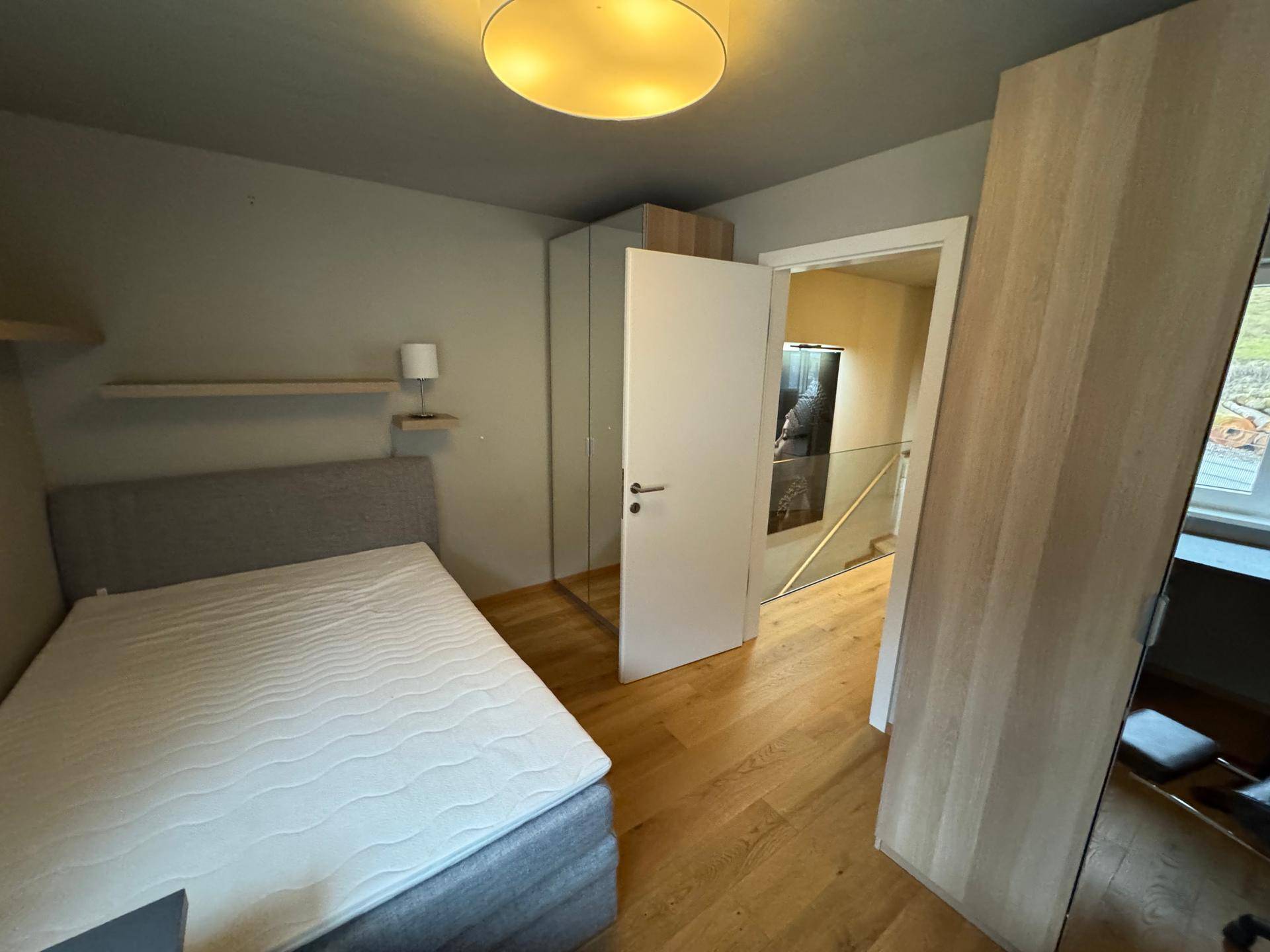 Helles Schlafzimmer mit Holzboden, schlichtem Bett und Zugang zu einem weiteren Bereich.