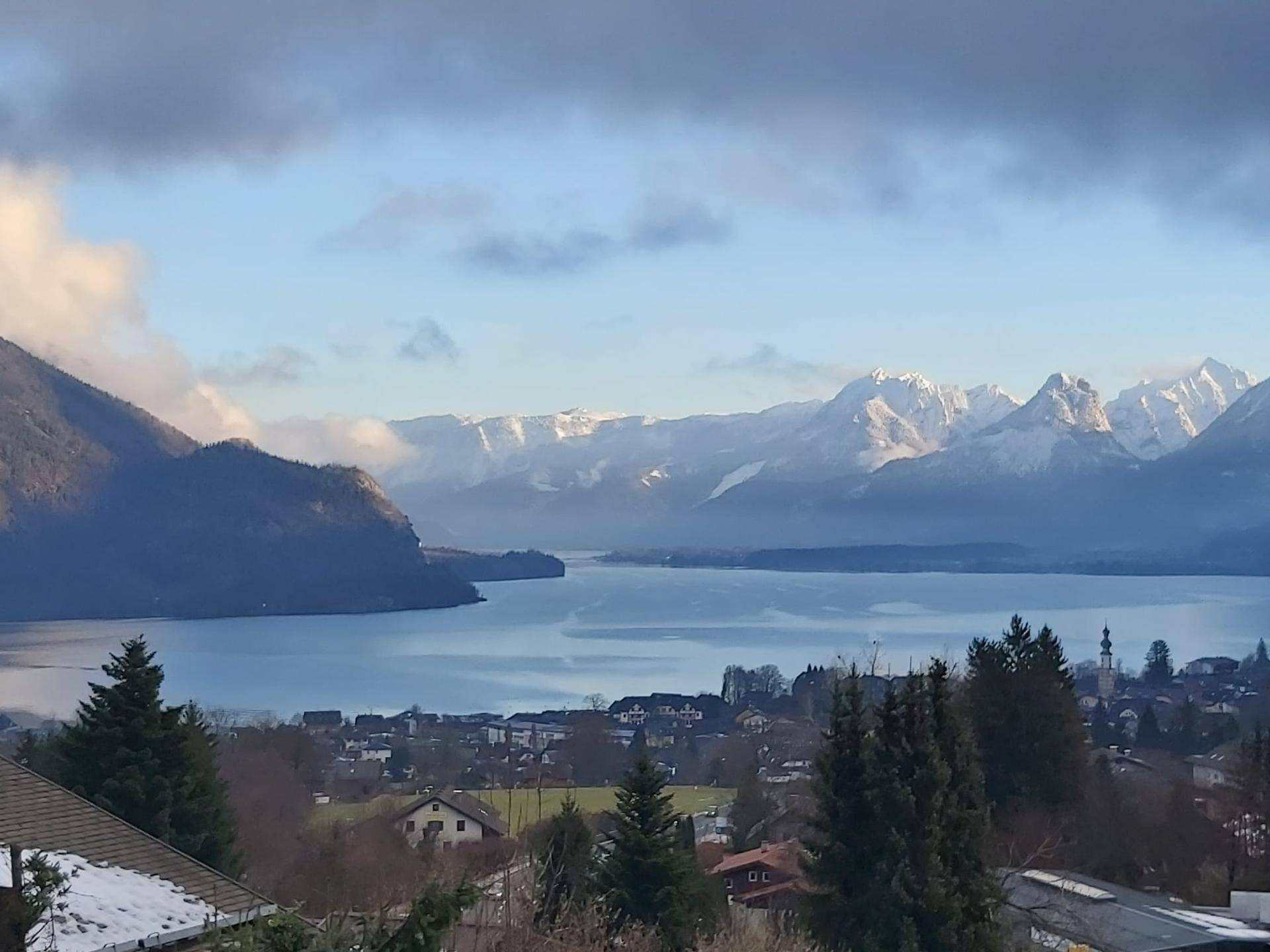 Panoramablick über einen See mit schneebedeckten Bergen und einer kleinen Ortschaft am Ufer.