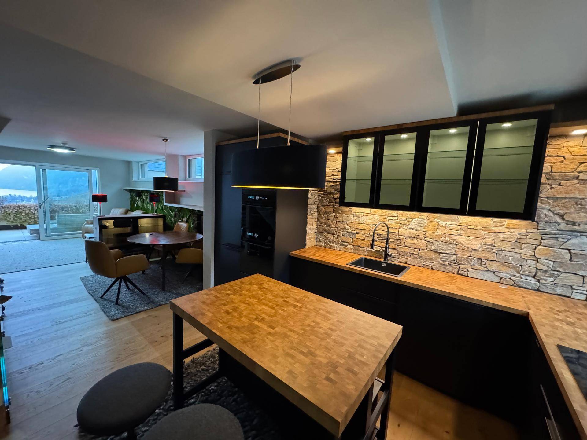 Moderne Küche mit schwarzer Einrichtung, Holztheke und Steinwand, offen zum Essbereich.