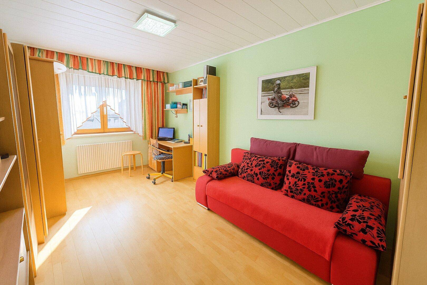 Helles Schlafzimmer mit grün gestrichenen Wänden, Schreibtisch und einem roten Sofa.
