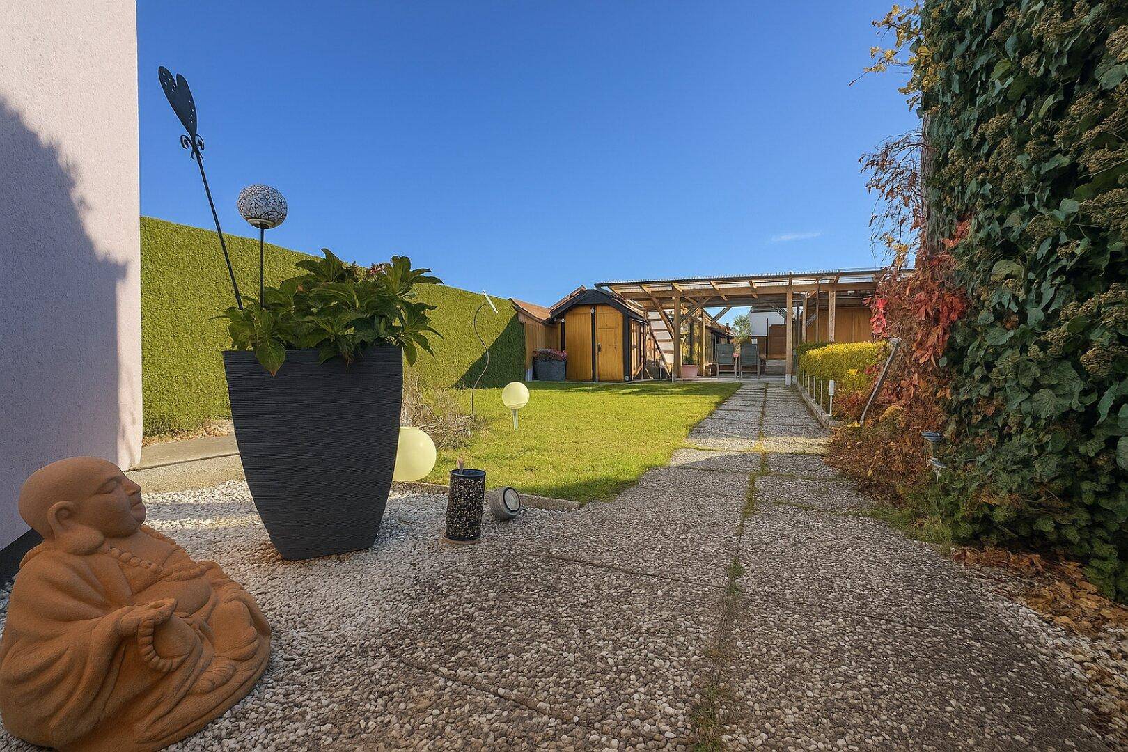 Gepflegter Garten mit Rasenfläche, Steinweg und einer Hecke im Hintergrund unter blauem Himmel.