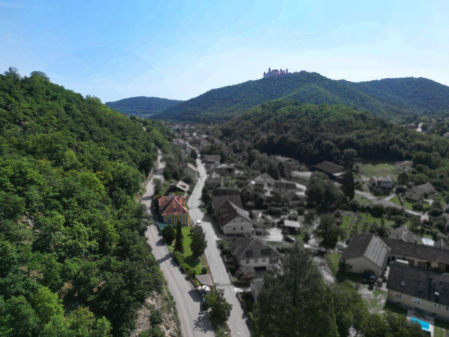 Weitläufige Luftaufnahme eines malerischen Dorfes in einem Tal, umgeben von grünen Hügeln und Wäldern.