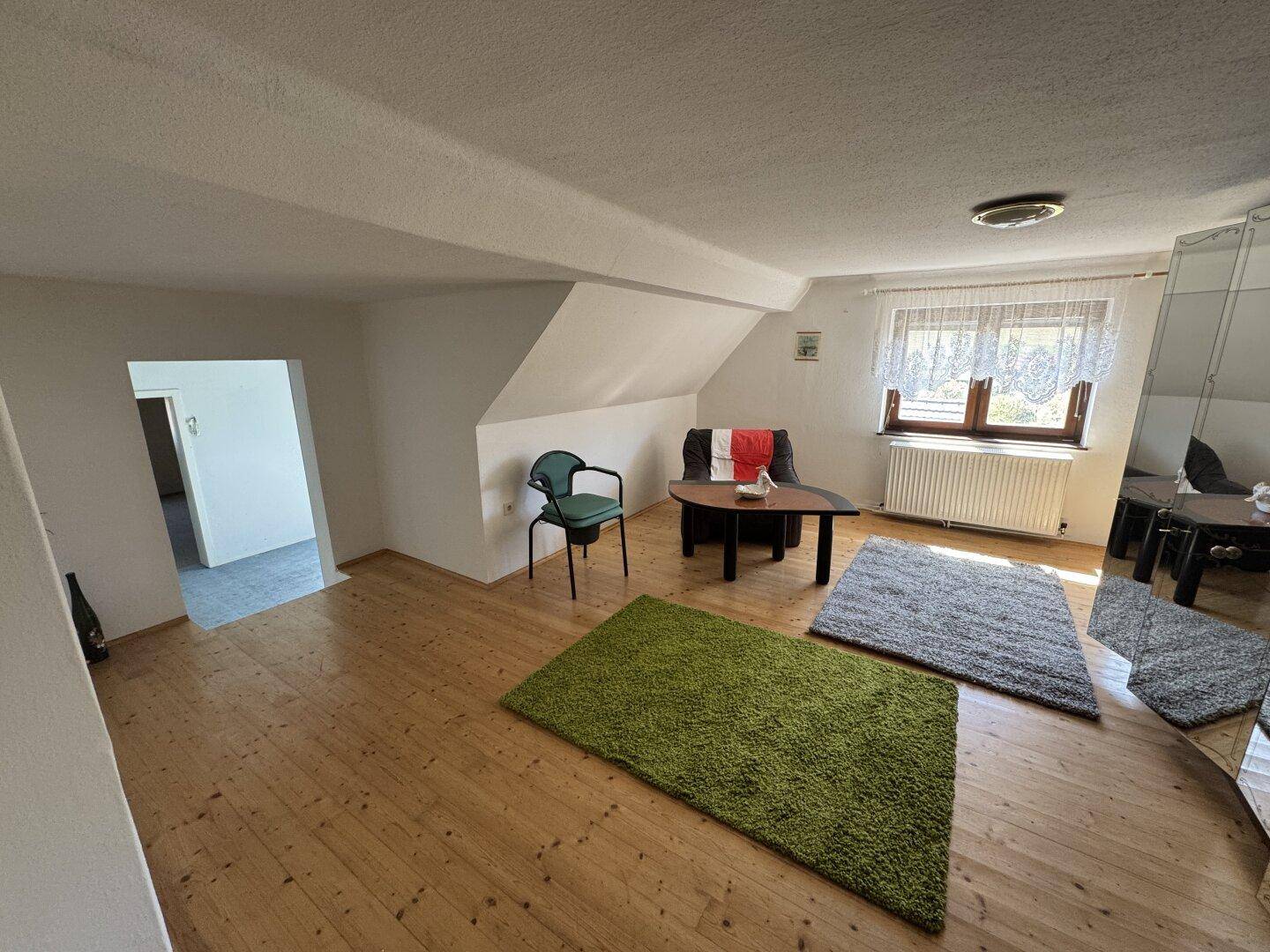 Geräumiges Dachzimmer mit Holzdielenboden, schrägen Decken und einfacher Einrichtung.