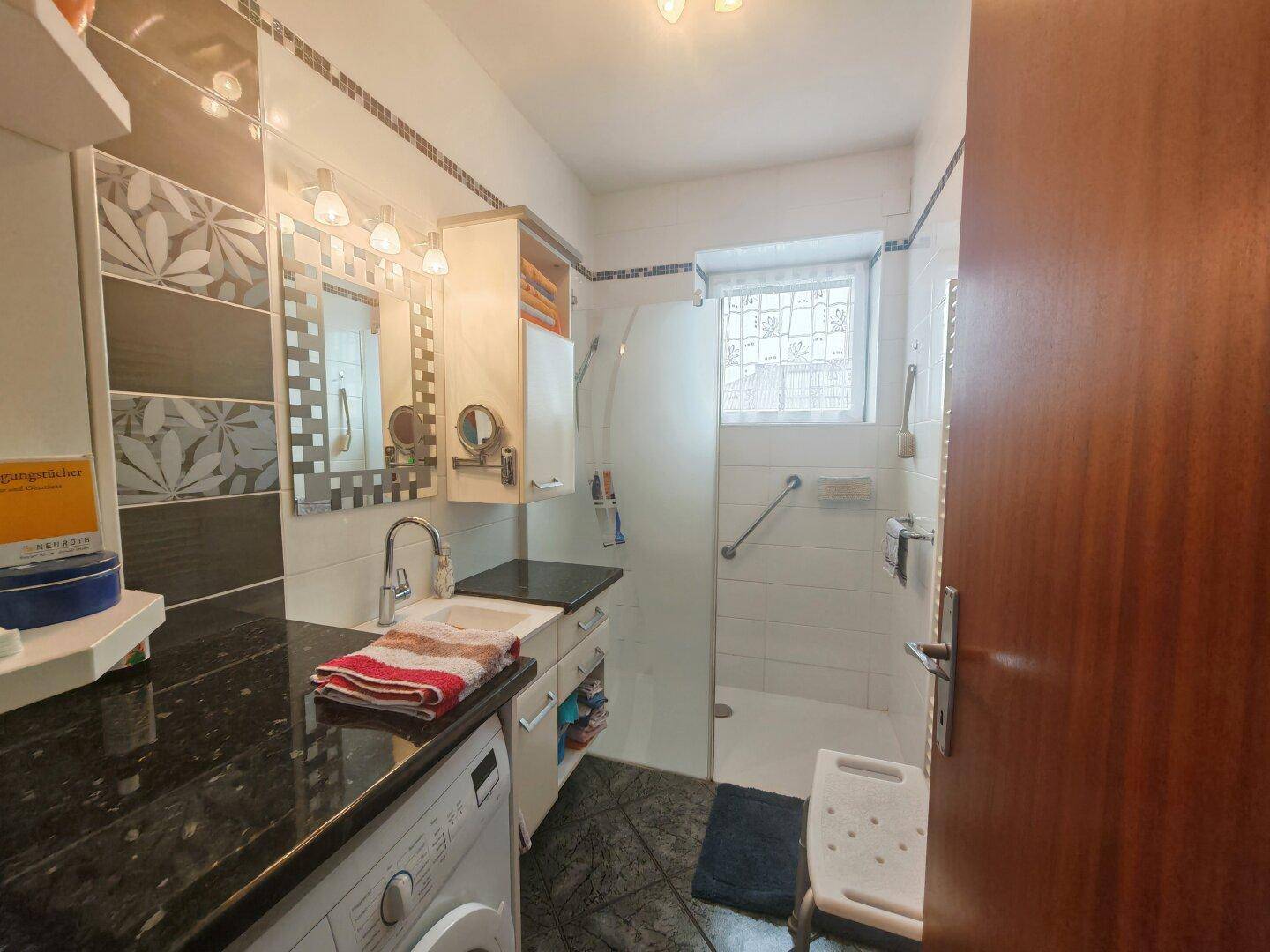 Modernes Badezimmer mit begehbarer Dusche, einzelnen Waschbecken, Haltegriffen und dunklen Fliesen.