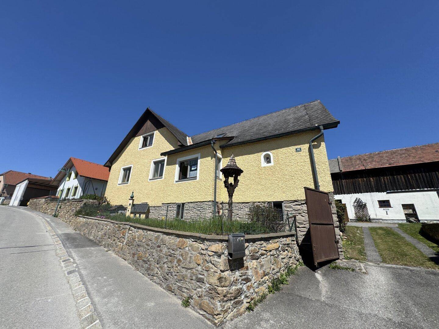 Gelbes Haus mit Satteldach und Steinmauer an einer leicht ansteigenden Straße unter blauem Himmel.