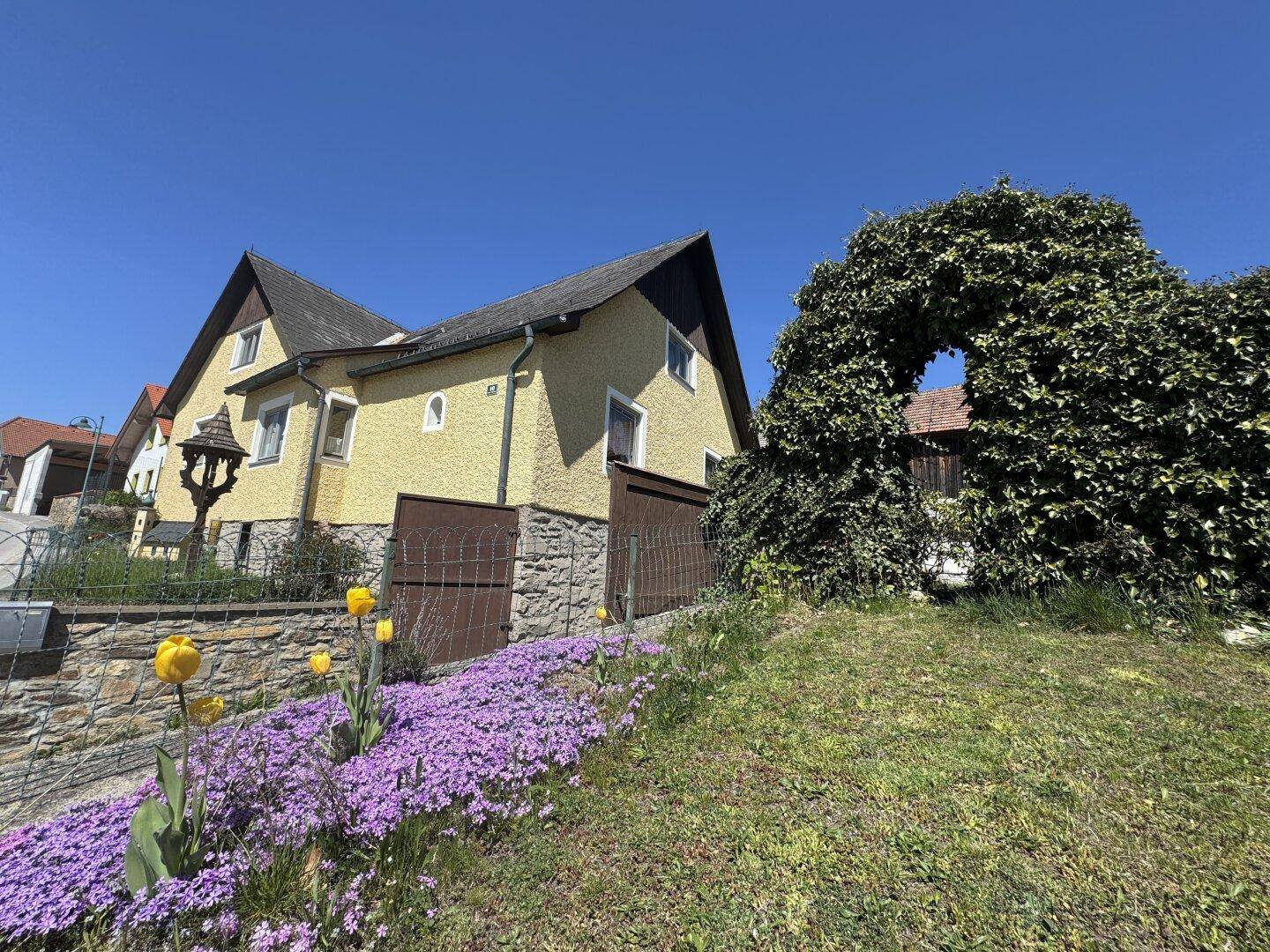 Gelbes Wohnhaus mit Satteldach, umgeben von blühenden Blumenbeeten und einem grünen Garten.