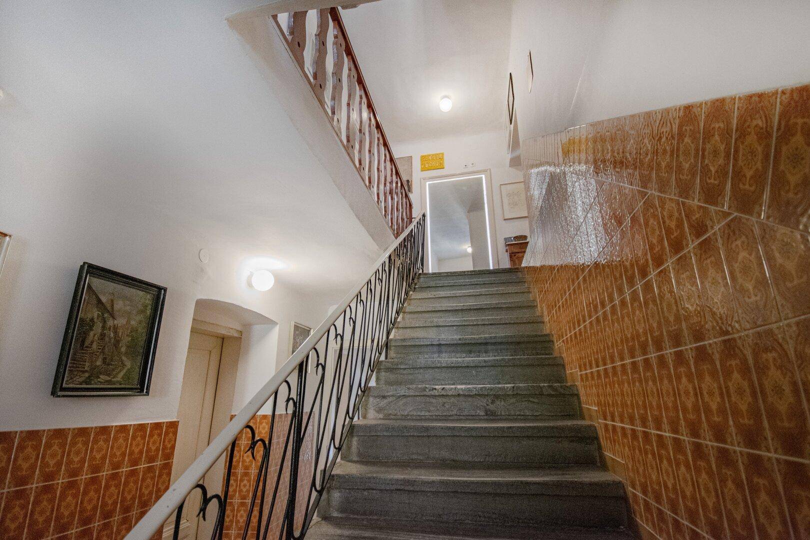 Treppenhaus mit gefliesten Wänden und schmiedeeisernem Geländer in einem älteren Gebäude.
