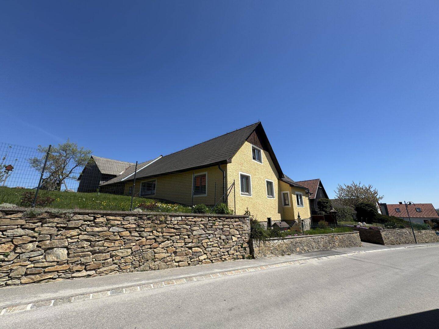 Gelbes Wohnhaus mit Satteldach und einer Natursteinmauer im Vordergrund unter strahlend blauem Himmel.