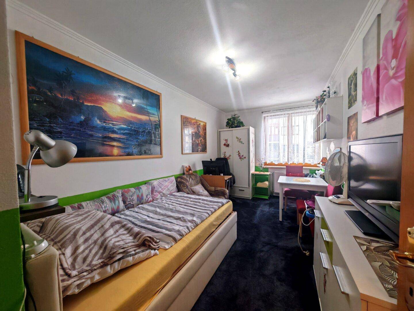 Schlafzimmer mit Bett, Schreibtisch und farbenfroher Wandgestaltung, hell durch großes Fenster.