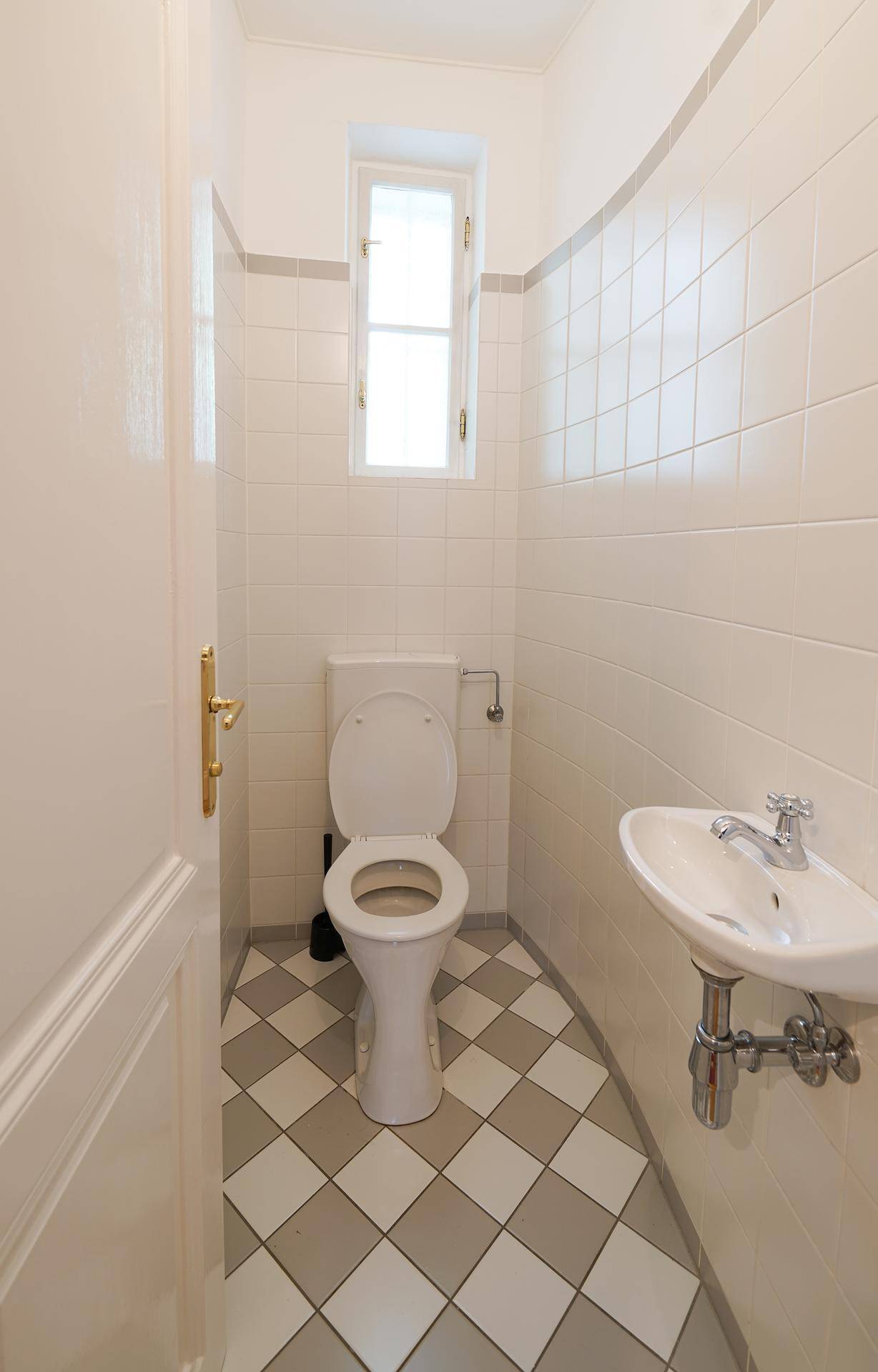 Kleines Gäste-WC mit Toilette, Waschbecken, Fenster und einem Schachbrettmuster auf dem Fliesenboden.