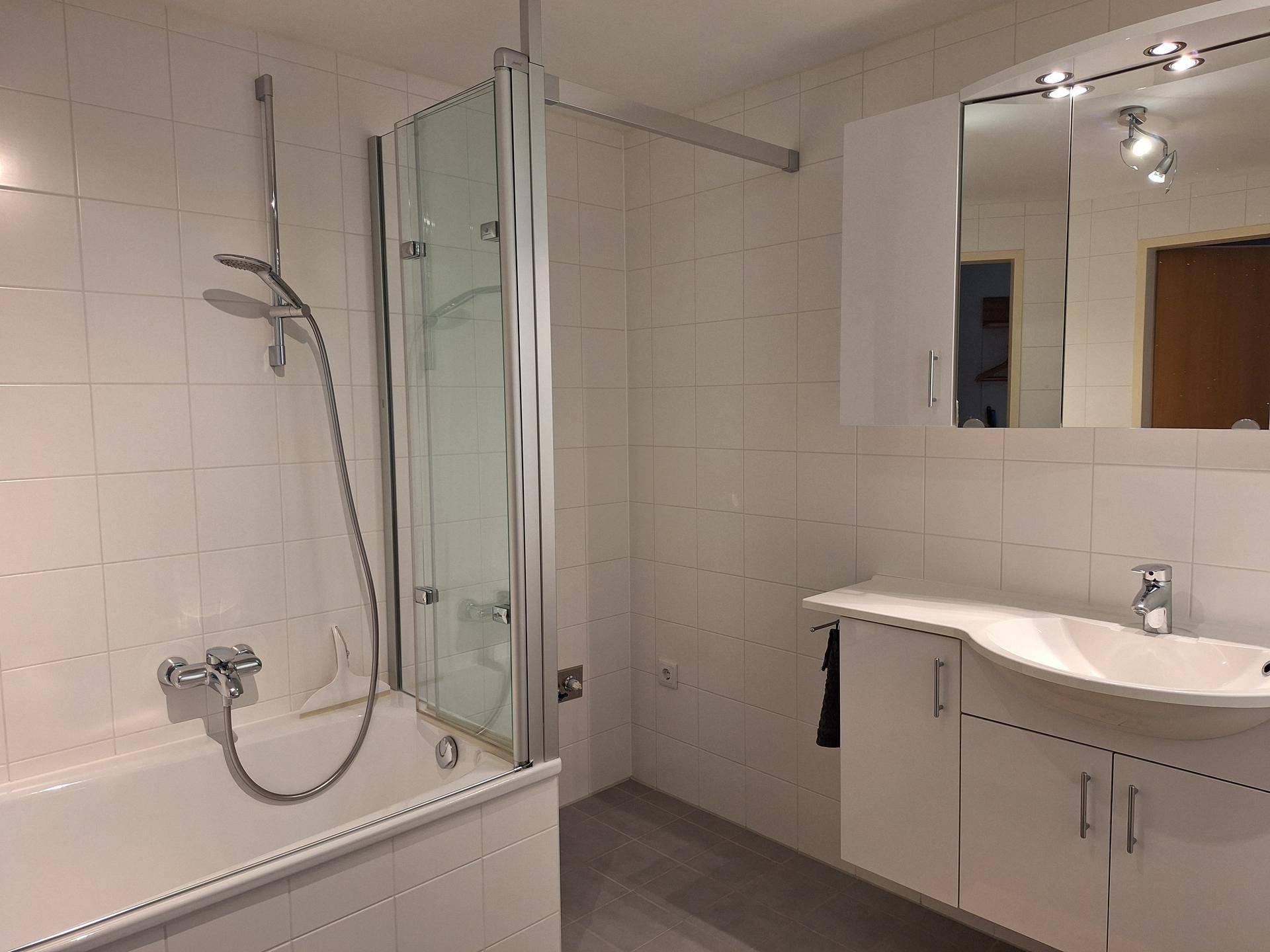 Helles Badezimmer mit weißen Fliesen, Badewanne mit Duschwand und einem Einzelwaschbecken mit Spiegelschrank.