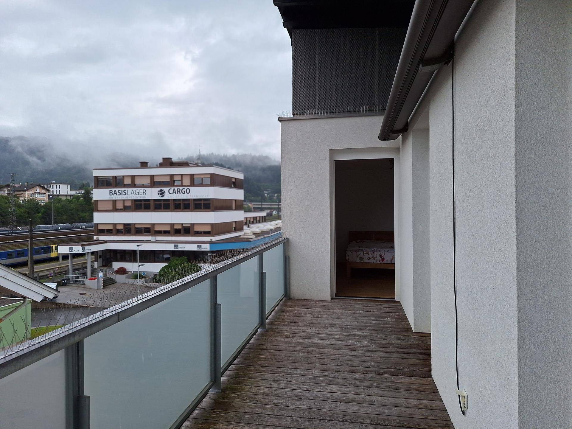Geräumiger Balkon mit Holzboden, Glasgeländer und Blick auf die Umgebung und die Berge im Hintergrund.