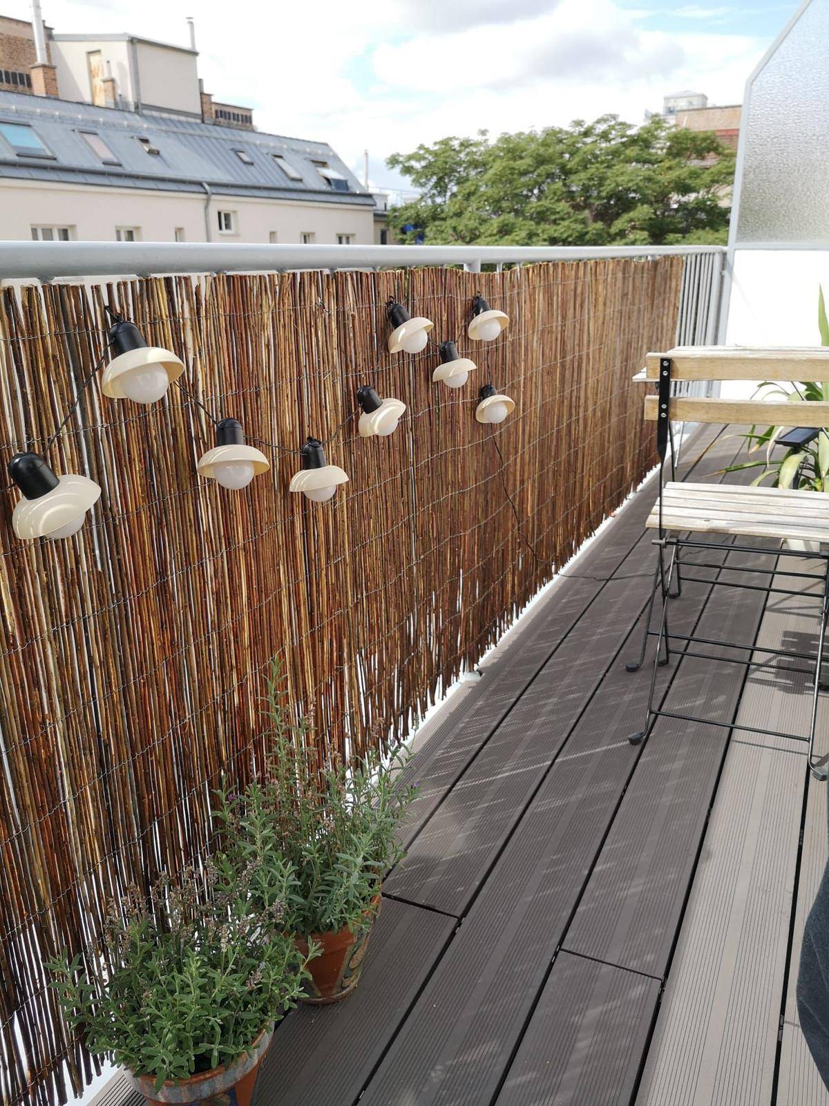 Gemütlicher Balkon mit Holzdielen, Bambus-Sichtschutz und dekorativer Lichterkette.