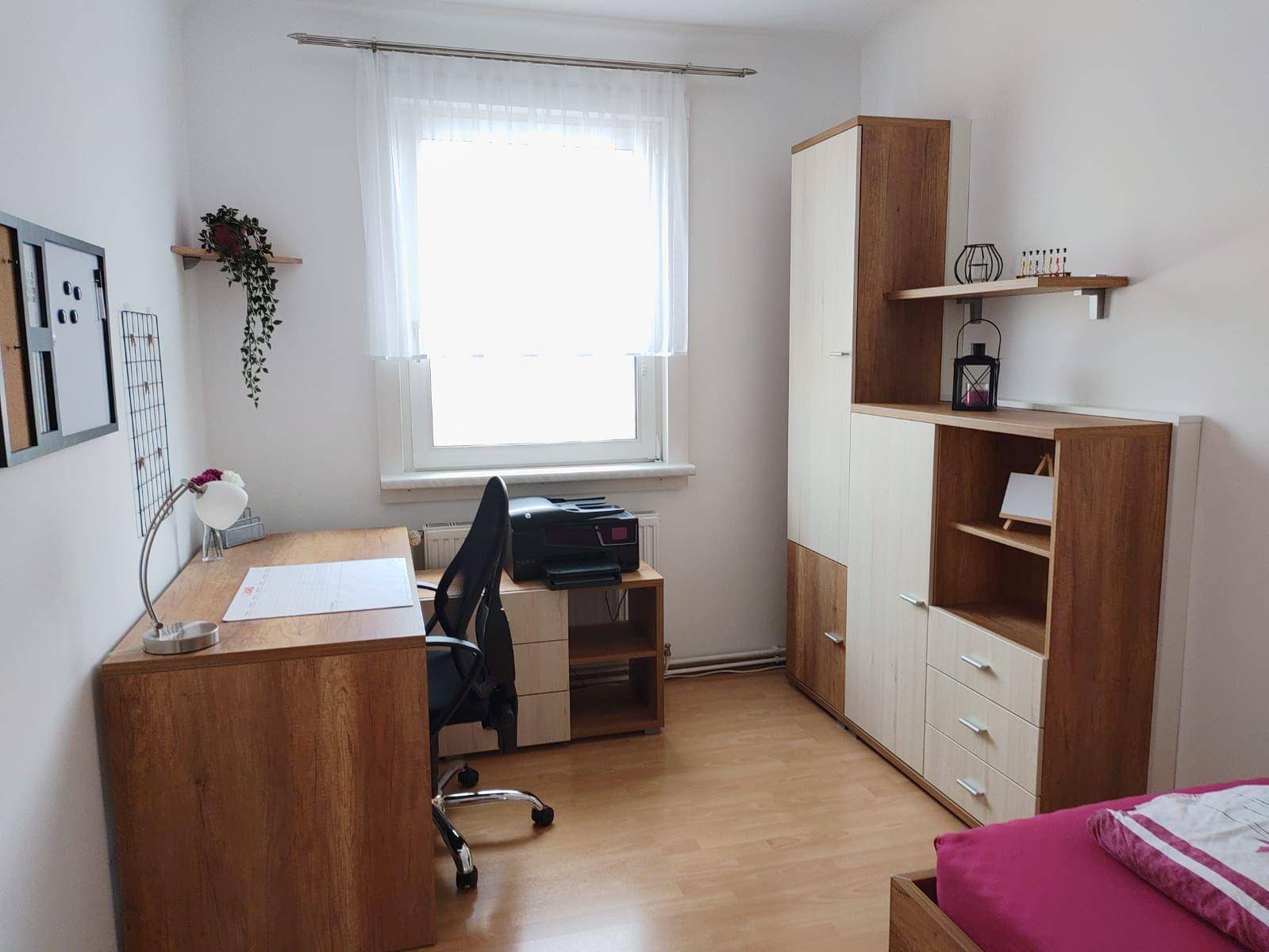 Funktionales Schlafzimmer mit Schreibtisch, Holzschrank und Fenster für natürliches Licht.