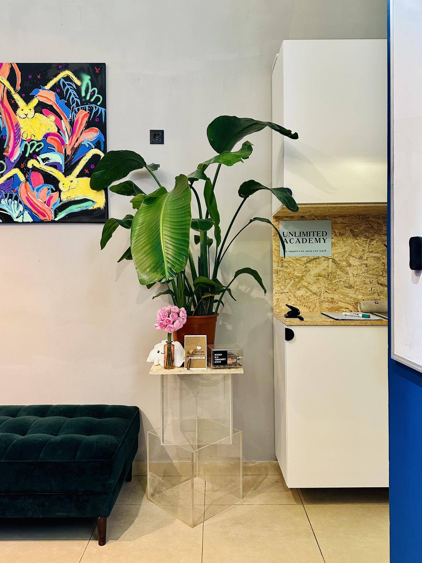Heller Bürobereich mit grüner Sitzbank, großer Zimmerpflanze und farbenfrohem Kunstwerk an der Wand.
