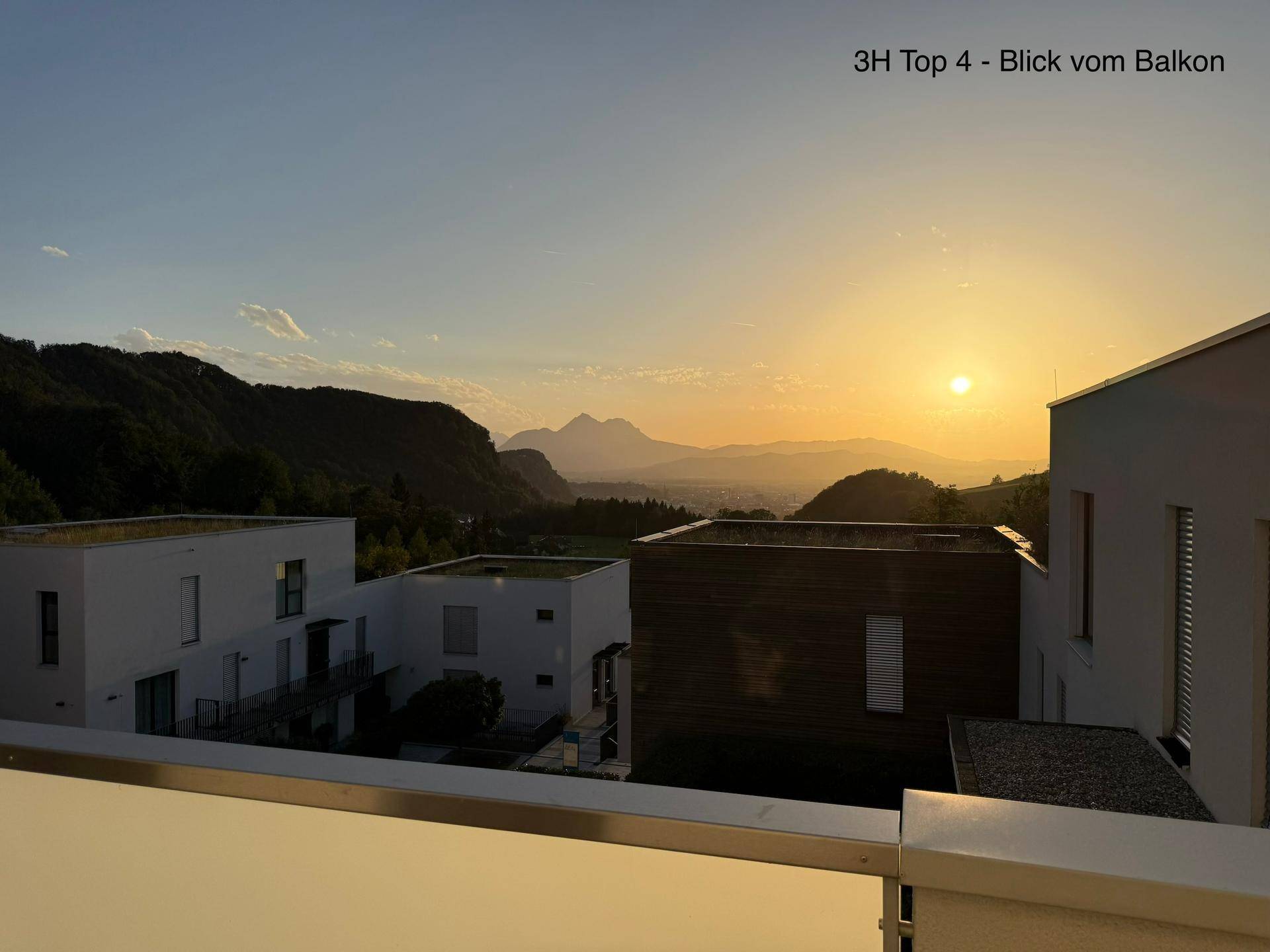 Panoramablick vom Balkon bei Sonnenuntergang auf moderne Gebäude, Hügel und ferne Berge.