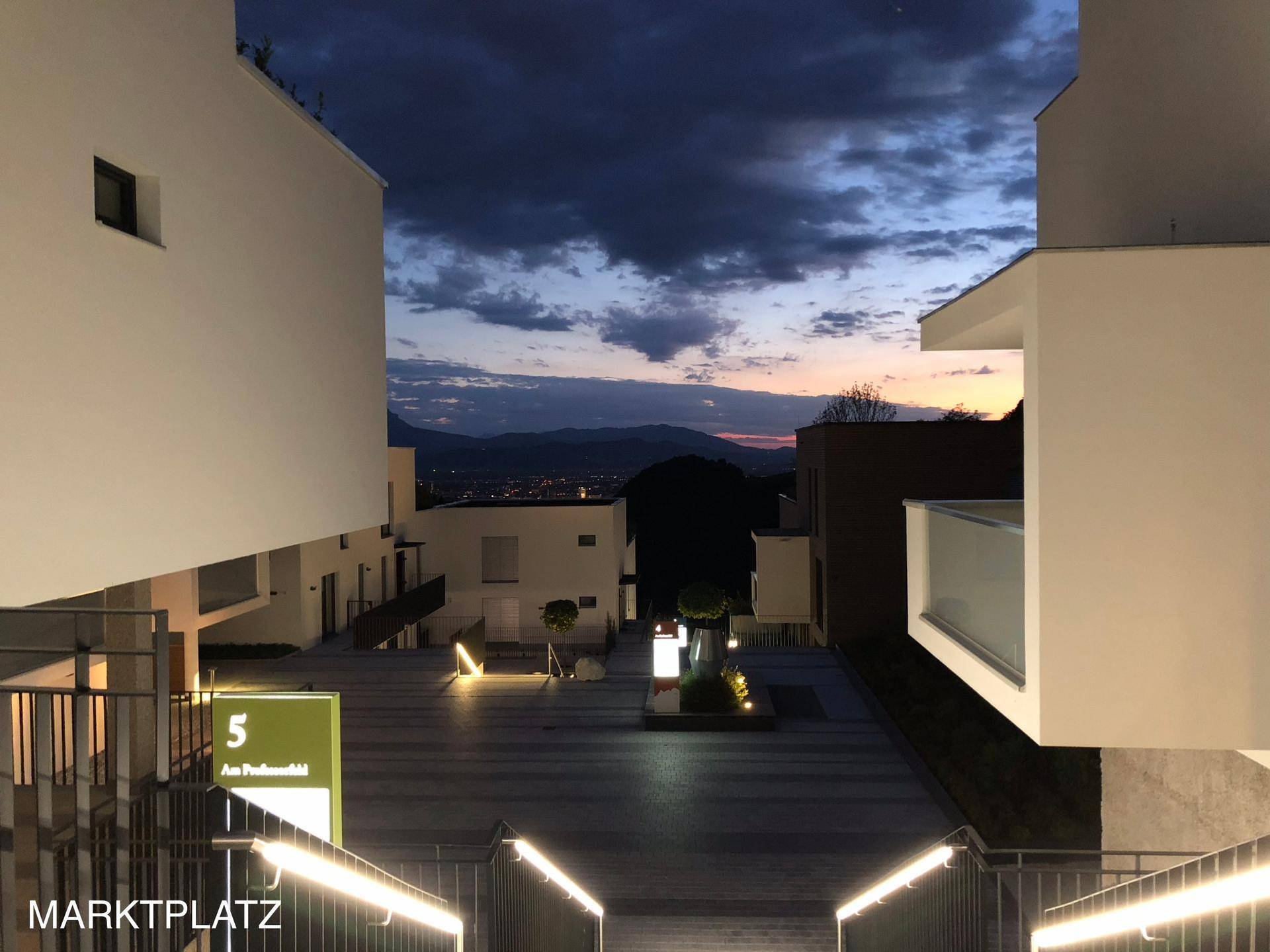 Beleuchteter moderner Innenhof zwischen weißen Gebäuden bei Sonnenuntergang mit Stadtblick in der Ferne.