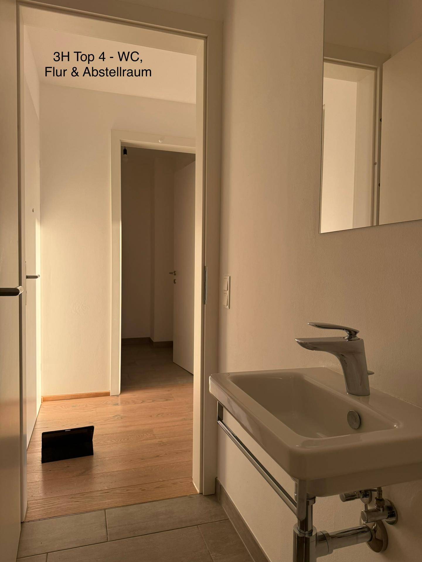 Blick aus einem WC in einen hellen Flur und einen Abstellraum mit Holzboden.