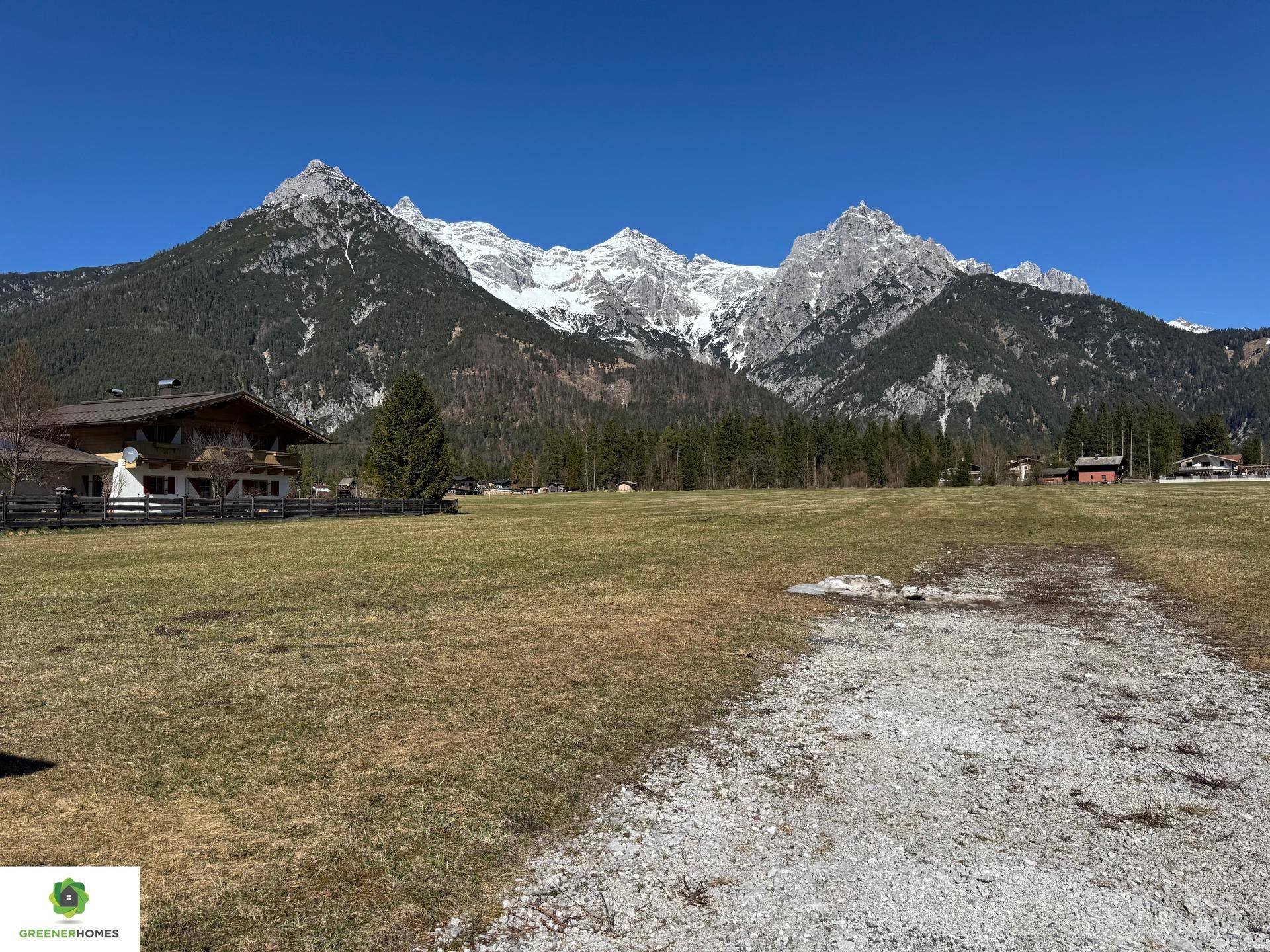 Idyllische Landschaft mit weitem Feld, traditionellen Gebäuden und beeindruckender Bergkulisse unter blauem Himmel.