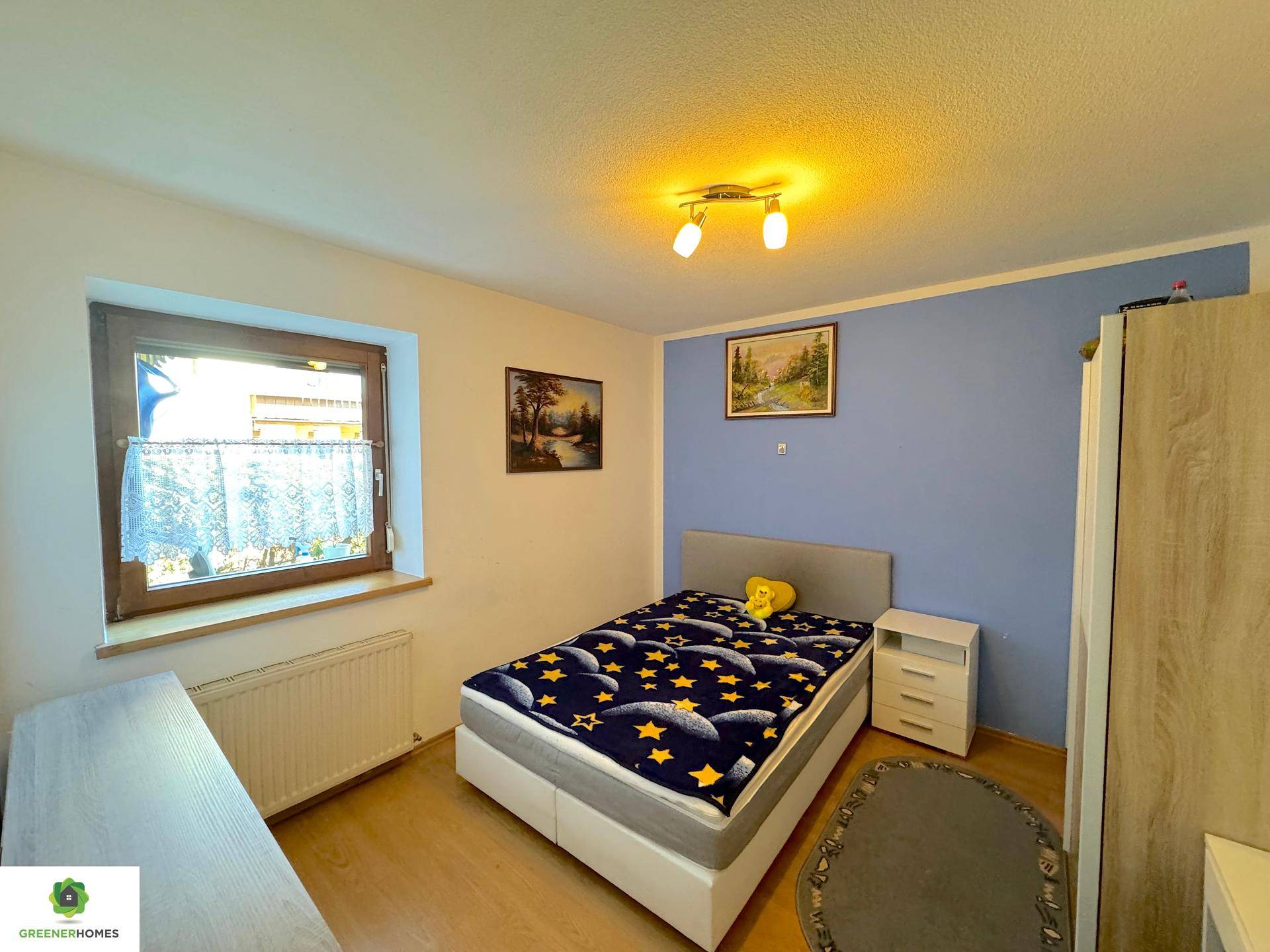 Schlafzimmer mit einer blauen Akzentwand, einem Bett und einem Fenster, das Tageslicht hereinlässt.