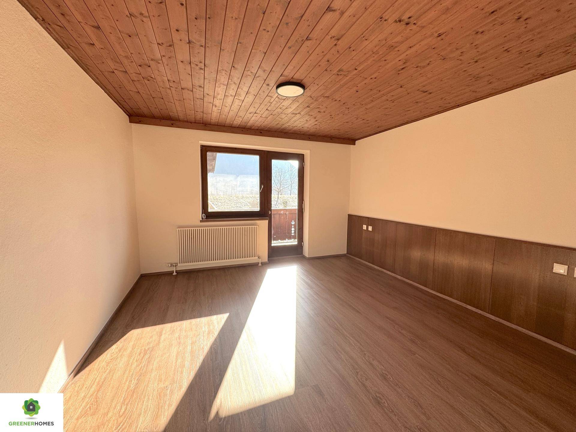 Helles Zimmer mit Holzbalkendecke, Laminatboden und Zugang zu einem kleinen Balkon.